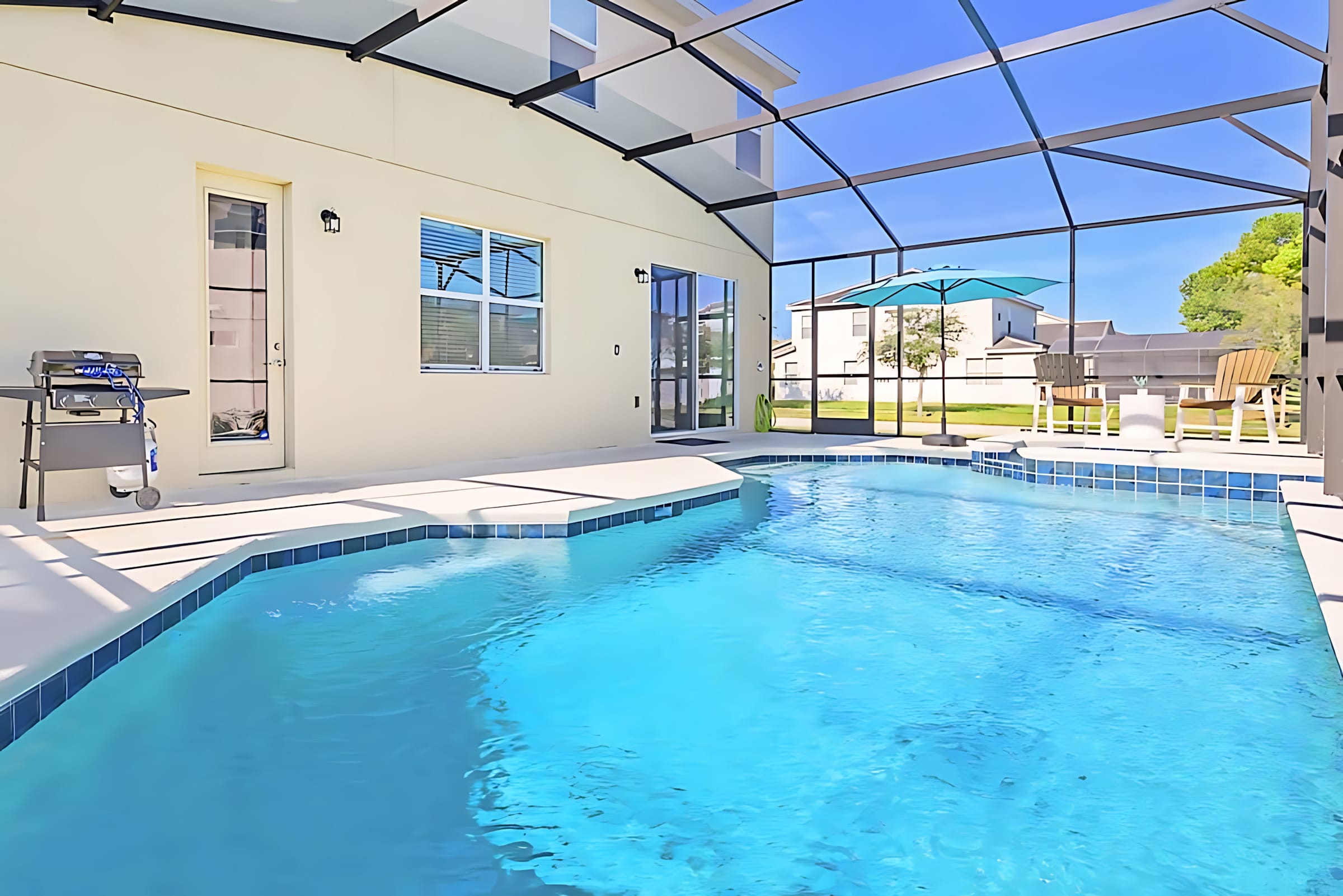 Wide view of a screened-in private pool area with a sparkling blue pool and hot tub, surrounded by a light-colored patio. The enclosure offers a clear view of the sunny sky and the exterior of the house.
