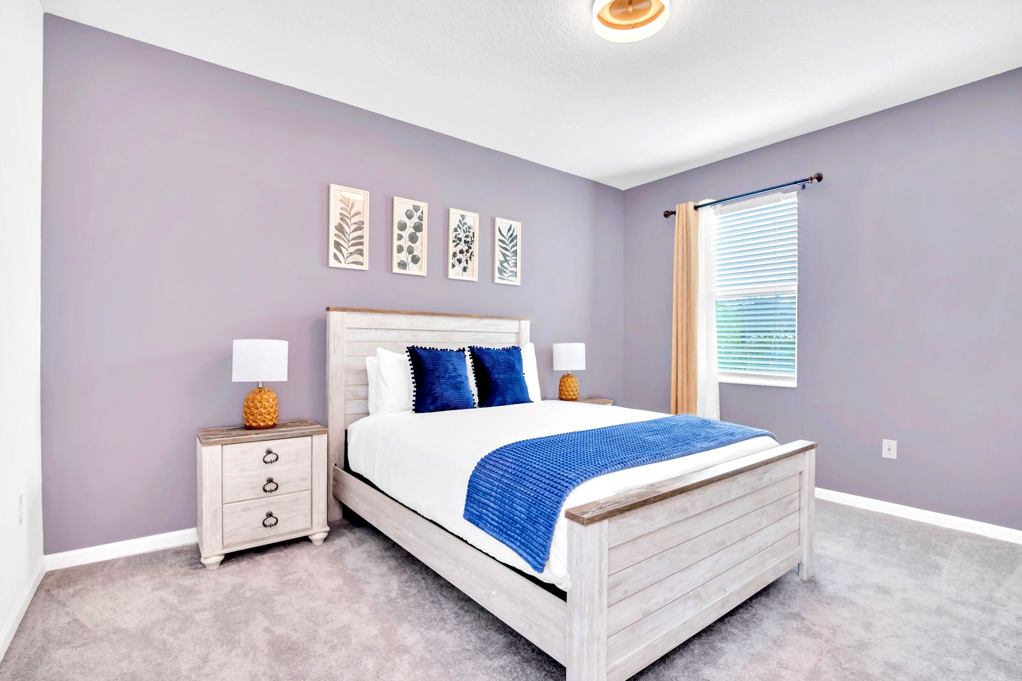 A spacious bedroom with light purple walls, light gray carpet, and a white distressed wooden bed frame with a white comforter and blue accents. Two matching nightstands with pineapple-shaped lamps flank the bed, and four botanical prints adorn the wall above the headboard.