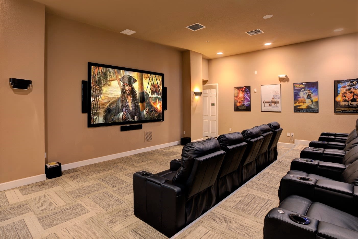 A dedicated home theater features a large projection screen displaying a movie scene, surrounded by multiple rows of black leather reclining seats with cup holders, set on a patterned carpet.