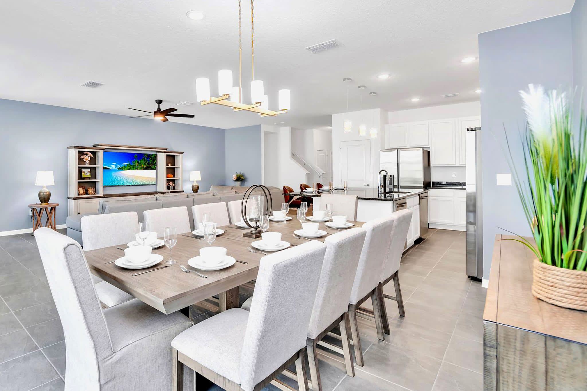 An expansive open-concept space showcases a dining area with a large wooden table and eight upholstered chairs, leading into a modern kitchen with white cabinetry and stainless steel appliances. The living room with a sectional sofa is visible in the background.