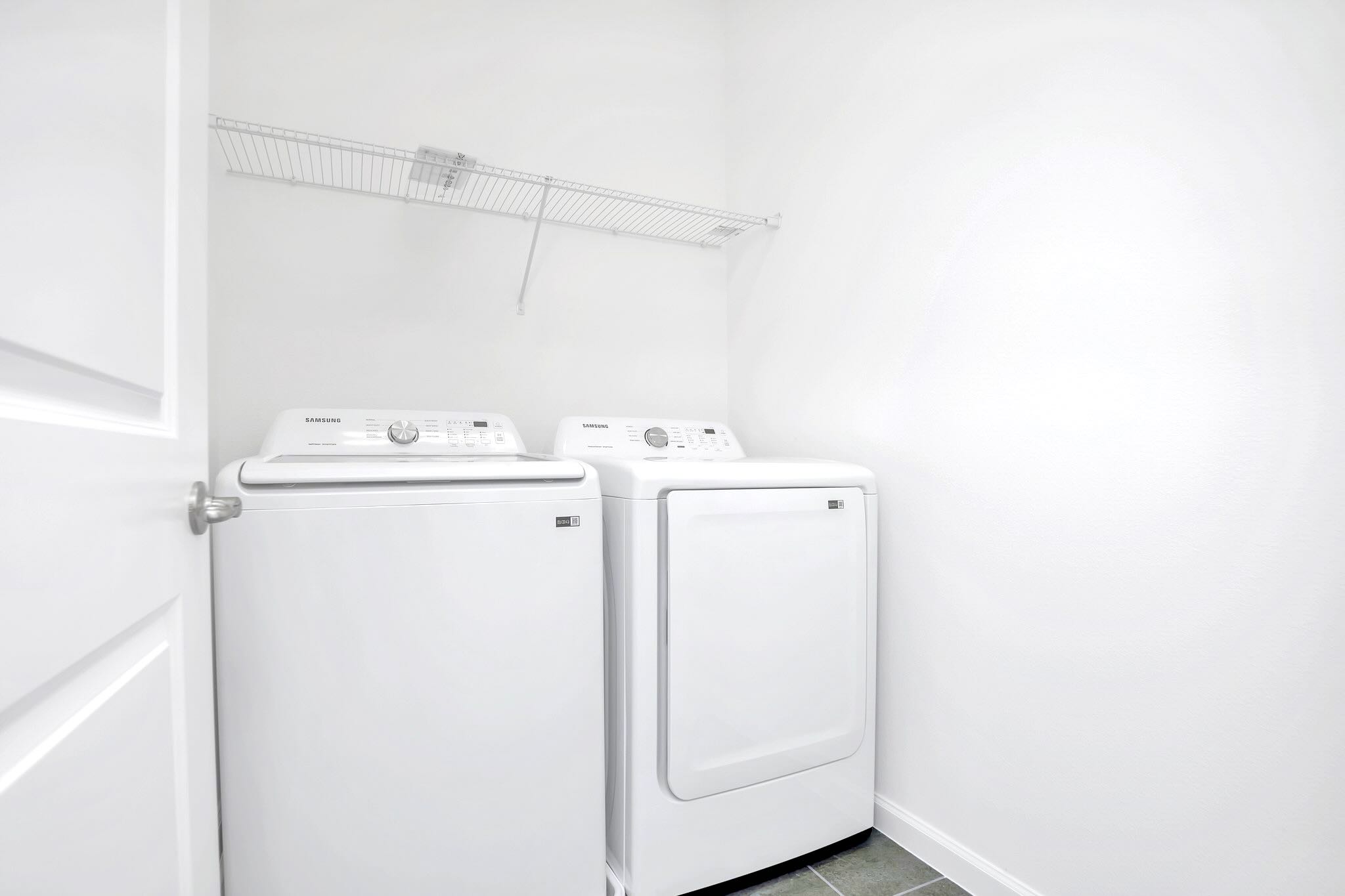 A functional laundry room with white walls and dark tiled floor, featuring a white Samsung top-load washing machine and a matching front-load dryer. A wire shelf is mounted above the appliances for storage.