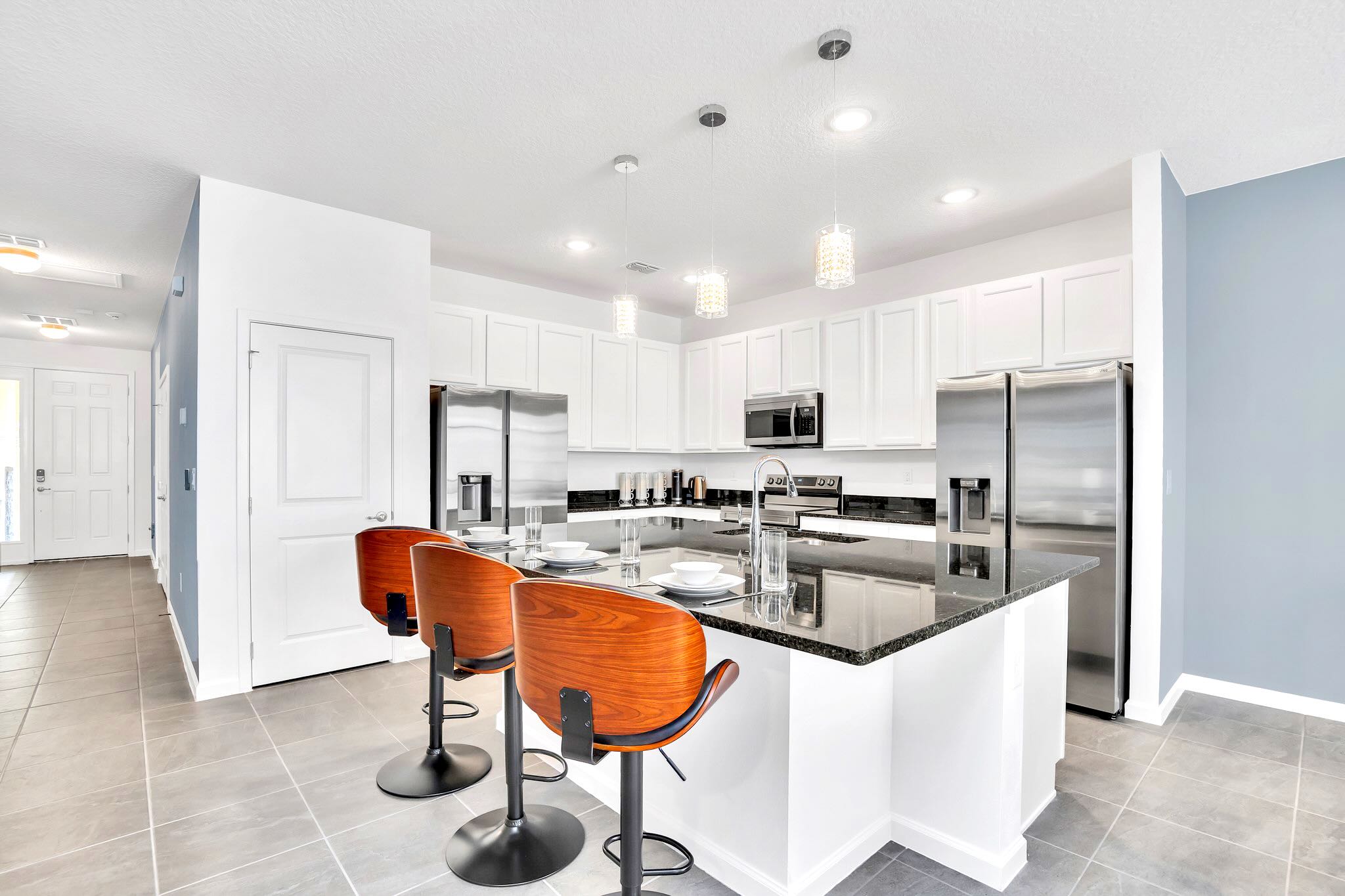 The bright kitchen features a large island with a sink and three wooden bar stools, set against white cabinetry and black granite countertops. Stainless steel appliances are visible, and the entrance to the home is seen in the background.