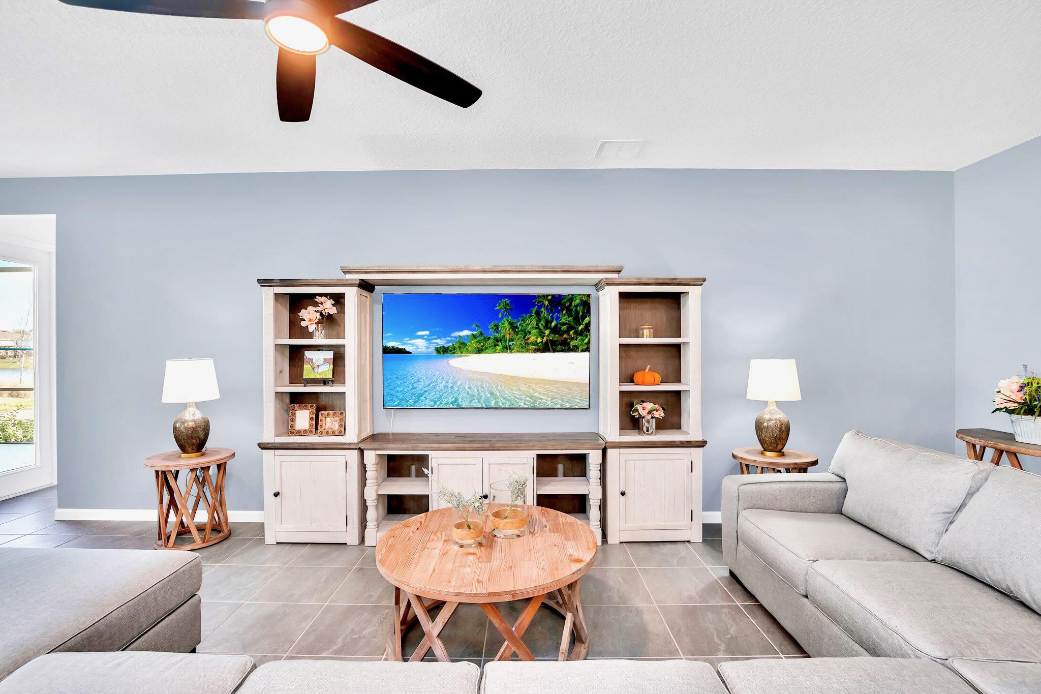 The living room features a large flat-screen TV mounted on a rustic white and wood entertainment center, flanked by two side tables with lamps. A round wooden coffee table sits in front of a light gray sectional sofa, all on light gray tiled flooring.