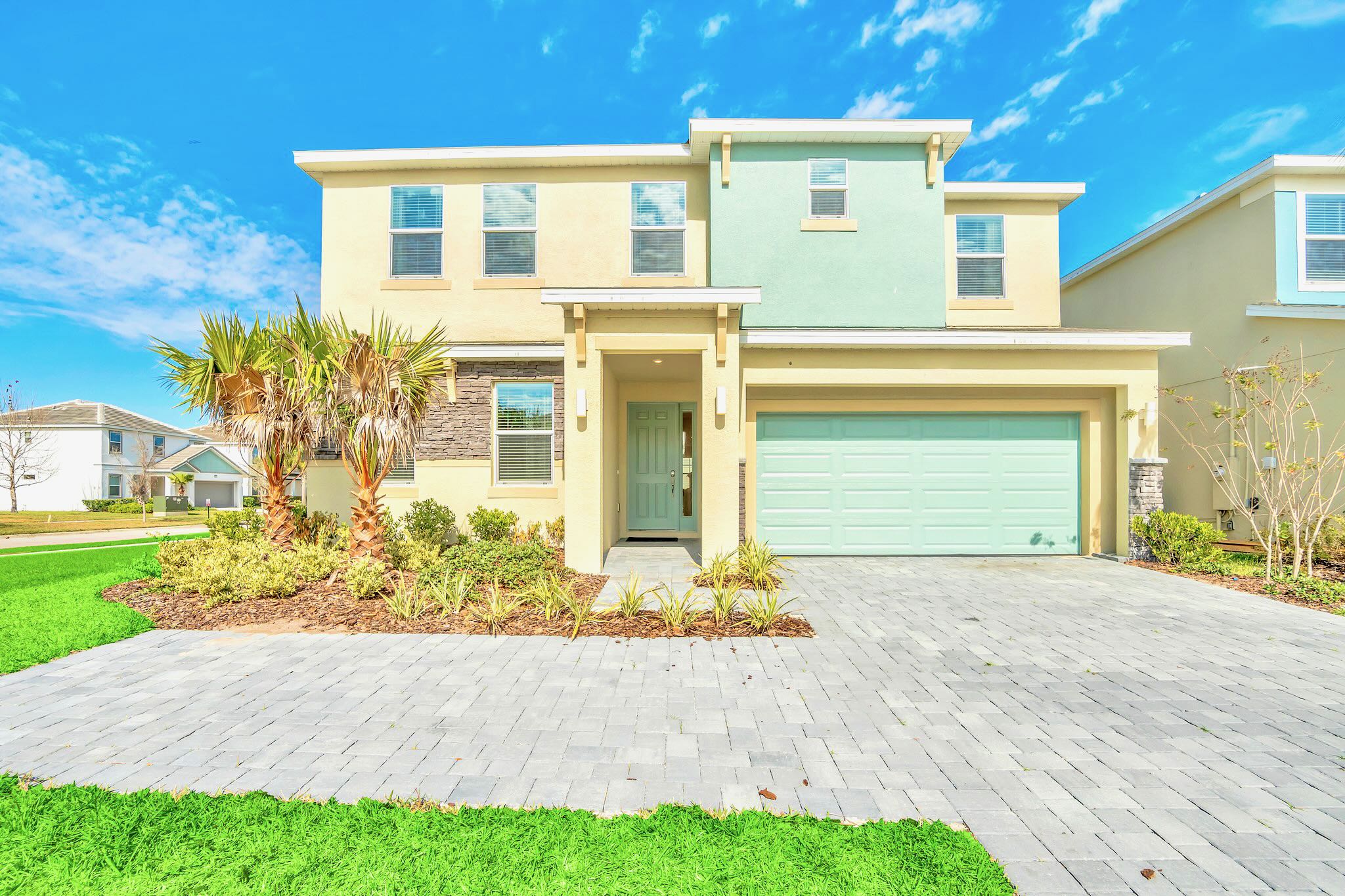 Modern two-story house with a light yellow and mint green facade, featuring a light blue front door and garage door, a paved driveway, and landscaped front yard with palm trees and green grass under a bright blue sky.