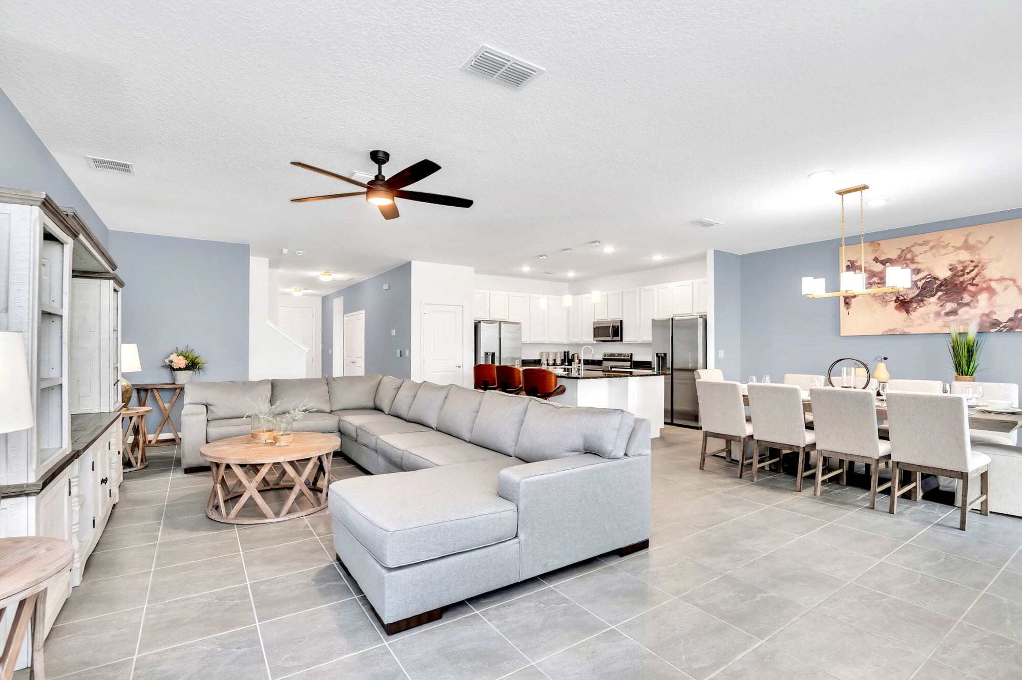A spacious open-concept living area features a large light gray sectional sofa around a round wooden coffee table, with a dining area and modern kitchen visible in the background. Light blue accent walls complement the light gray tiled floor and white ceilings.