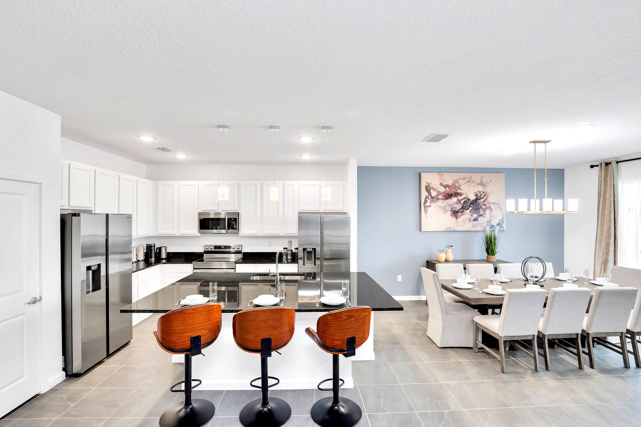 A modern kitchen features white shaker cabinets, black granite countertops, and stainless steel appliances, including two refrigerators. A large island with a sink offers seating for three on stylish wooden bar stools, illuminated by pendant lights.