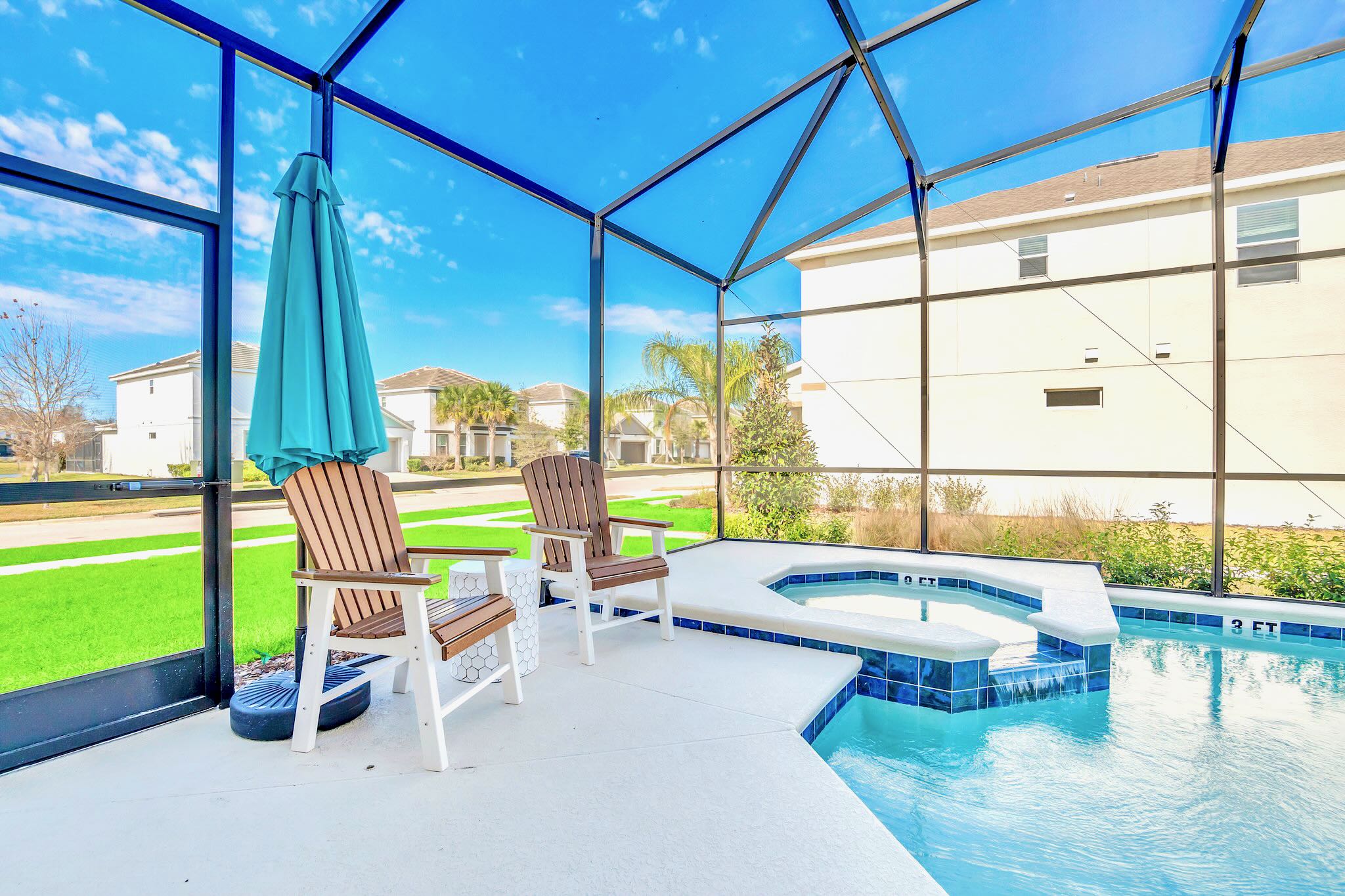 Screened-in private pool area with a hot tub in the foreground and a pool extending beyond, featuring two wooden Adirondack chairs and a turquoise umbrella on the patio. The enclosure provides views of a grassy area and neighboring houses.