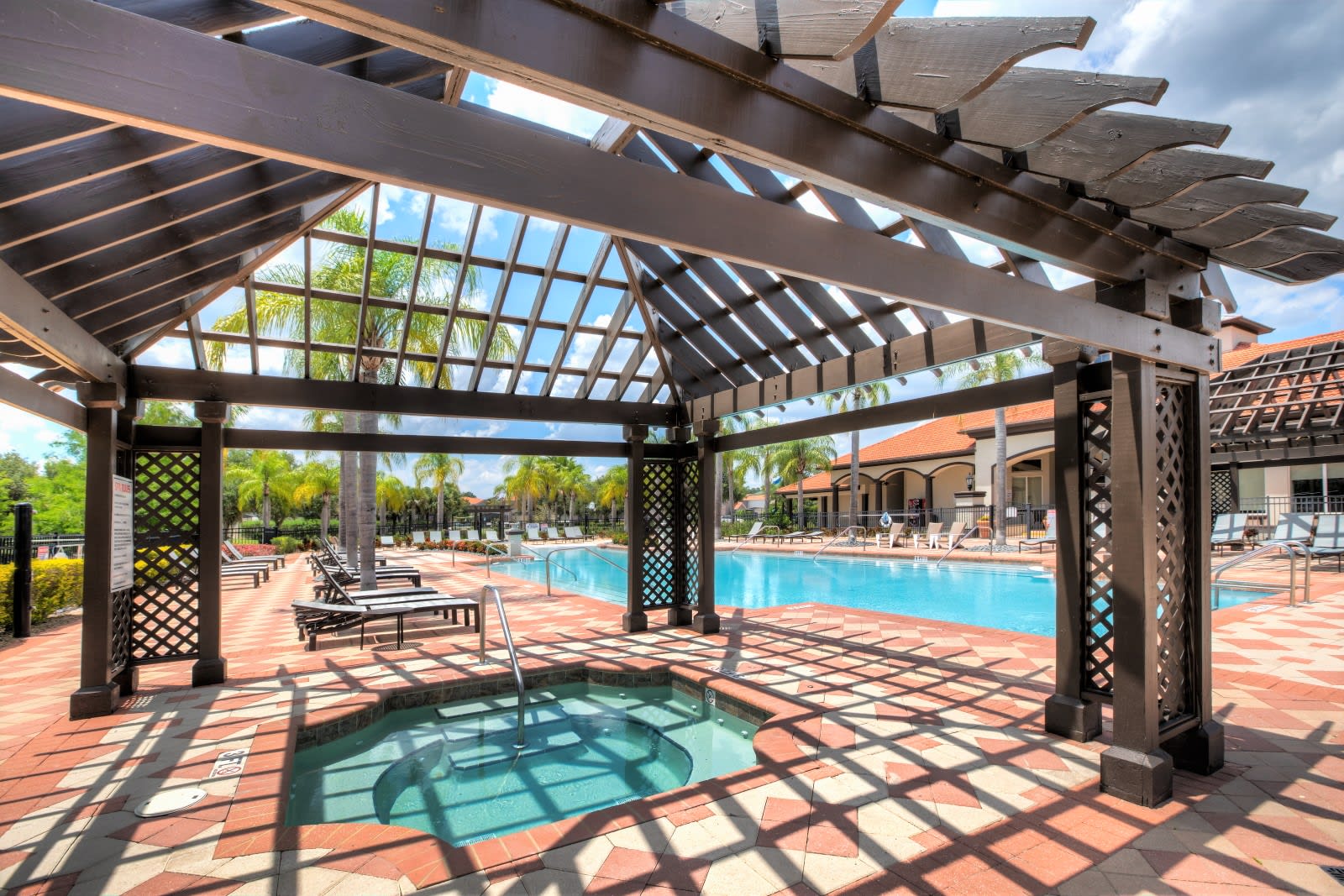 A community pool area features a built-in hot tub under a dark wooden pergola, with lounge chairs visible around the main swimming pool, all set on a patterned brick patio under a bright sky.