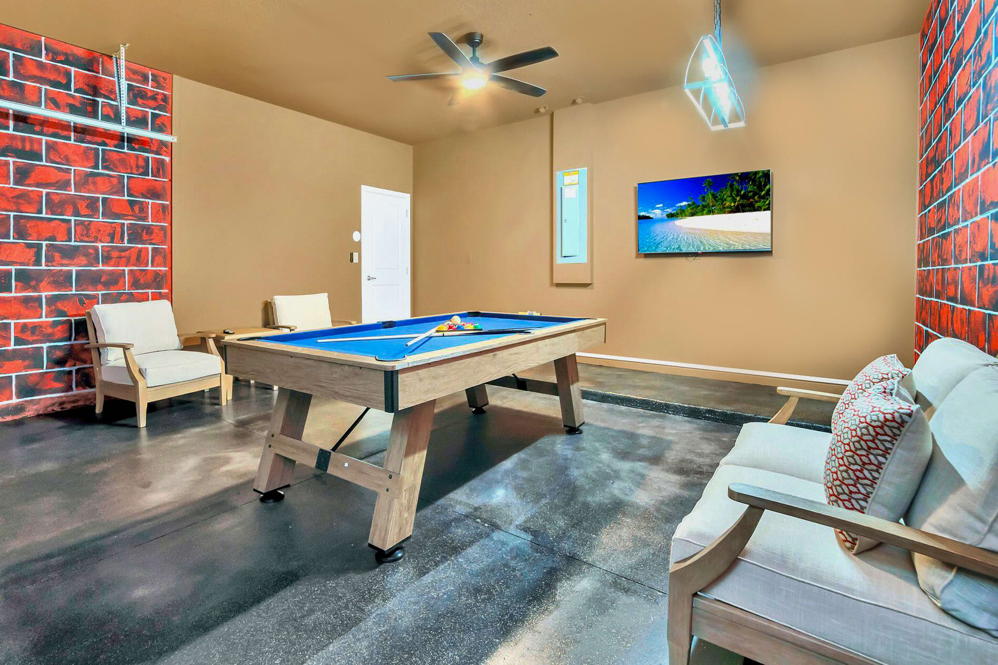 Game room with a pool table at its center, featuring exposed brick-patterned walls, a mounted flat-screen TV, and casual seating including a sofa and armchairs. The floor is a dark, polished concrete.