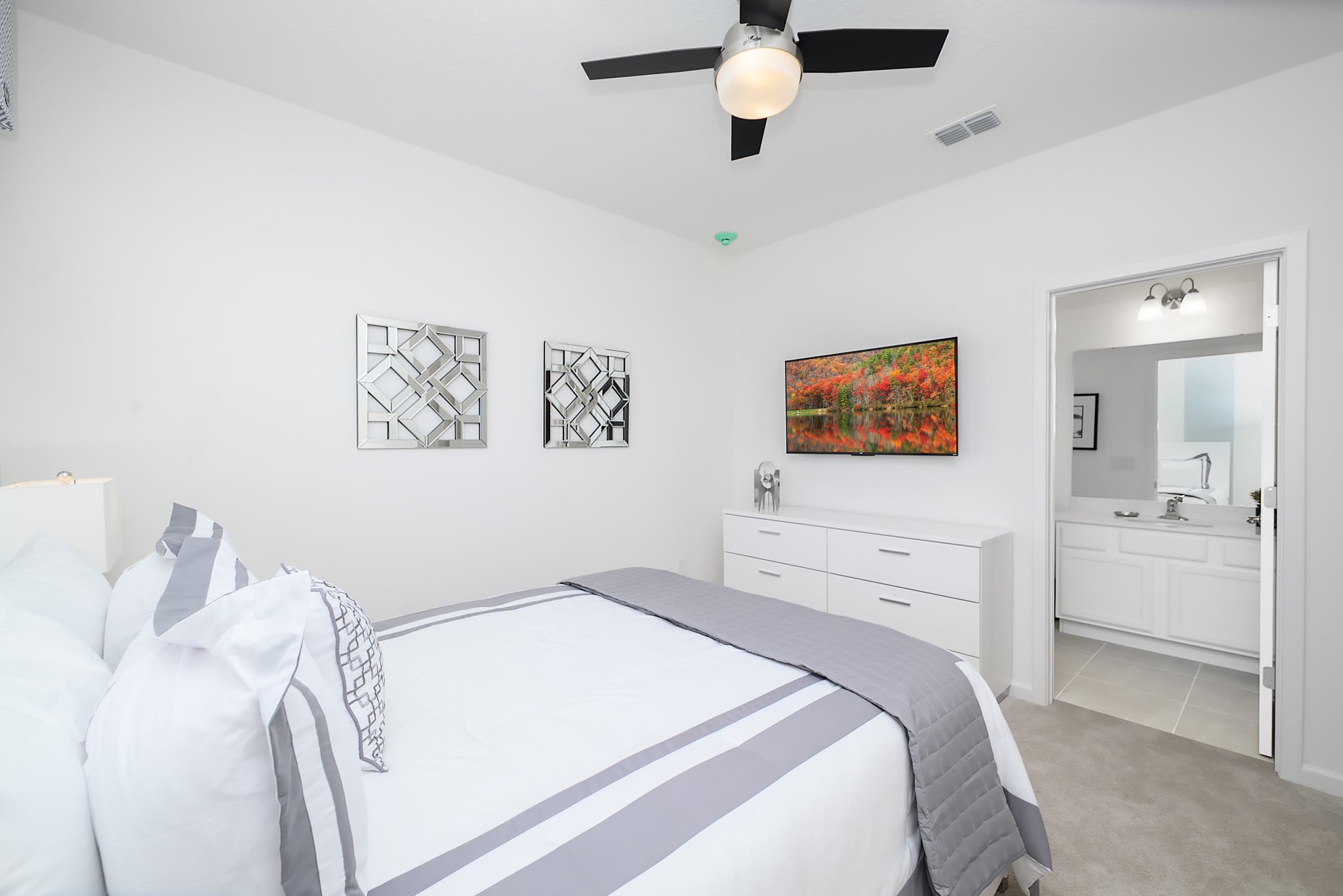 View of a modern bedroom with a white dresser and a wall-mounted TV, showing a glimpse into an en-suite bathroom with a white vanity and mirror. The room has light carpet and white walls, with decorative square mirrors on the wall.