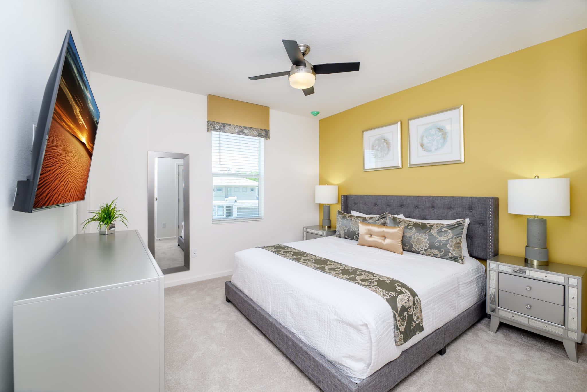 A vibrant bedroom with a king-sized bed featuring a gray upholstered headboard and a decorative throw. A yellow accent wall behind the bed is adorned with two framed artworks, and a ceiling fan hangs overhead.