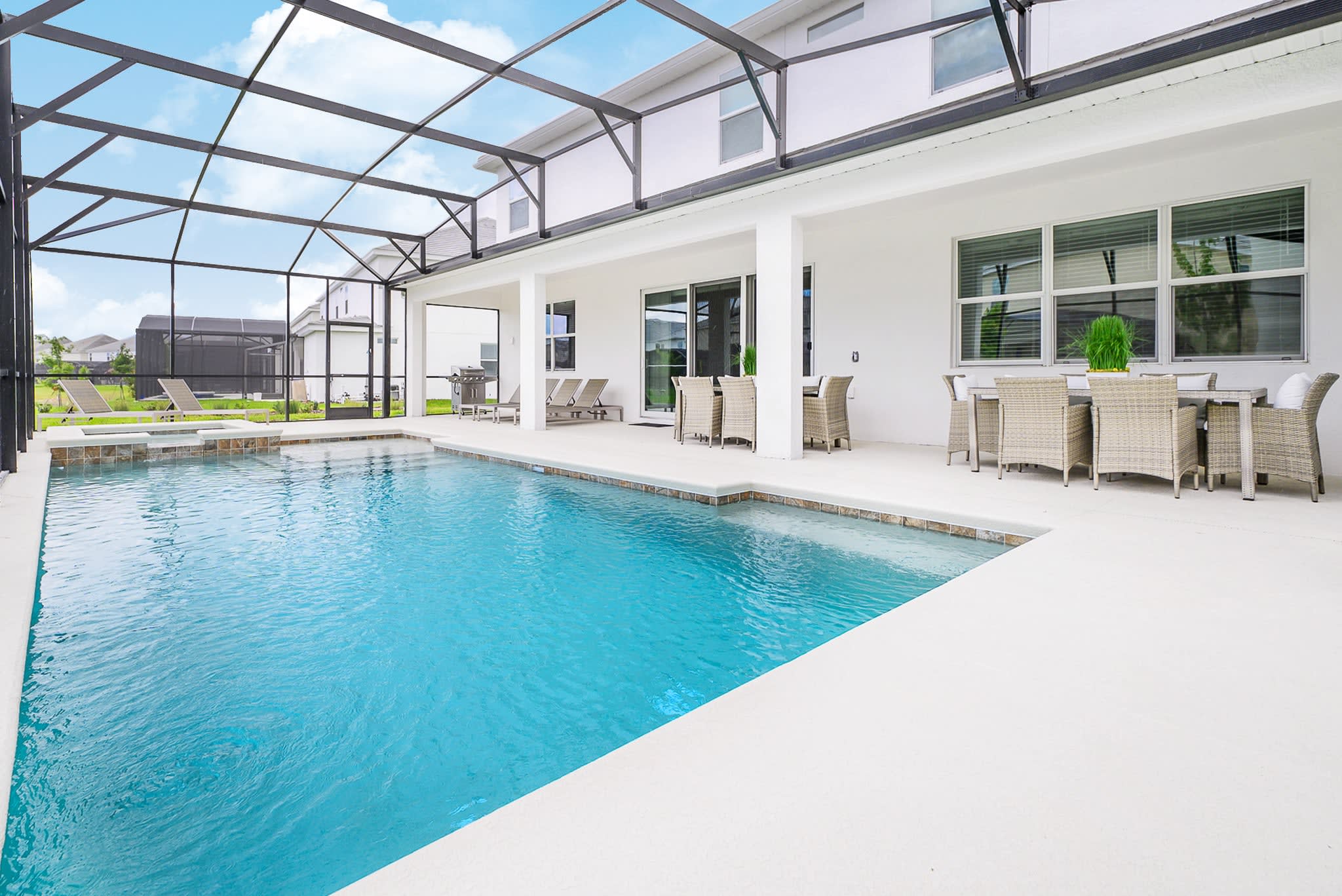 A wide view of the screened-in outdoor pool area, showcasing a large swimming pool and integrated hot tubs. The white patio is furnished with lounge chairs, and the house exterior features multiple windows and sliding glass doors.