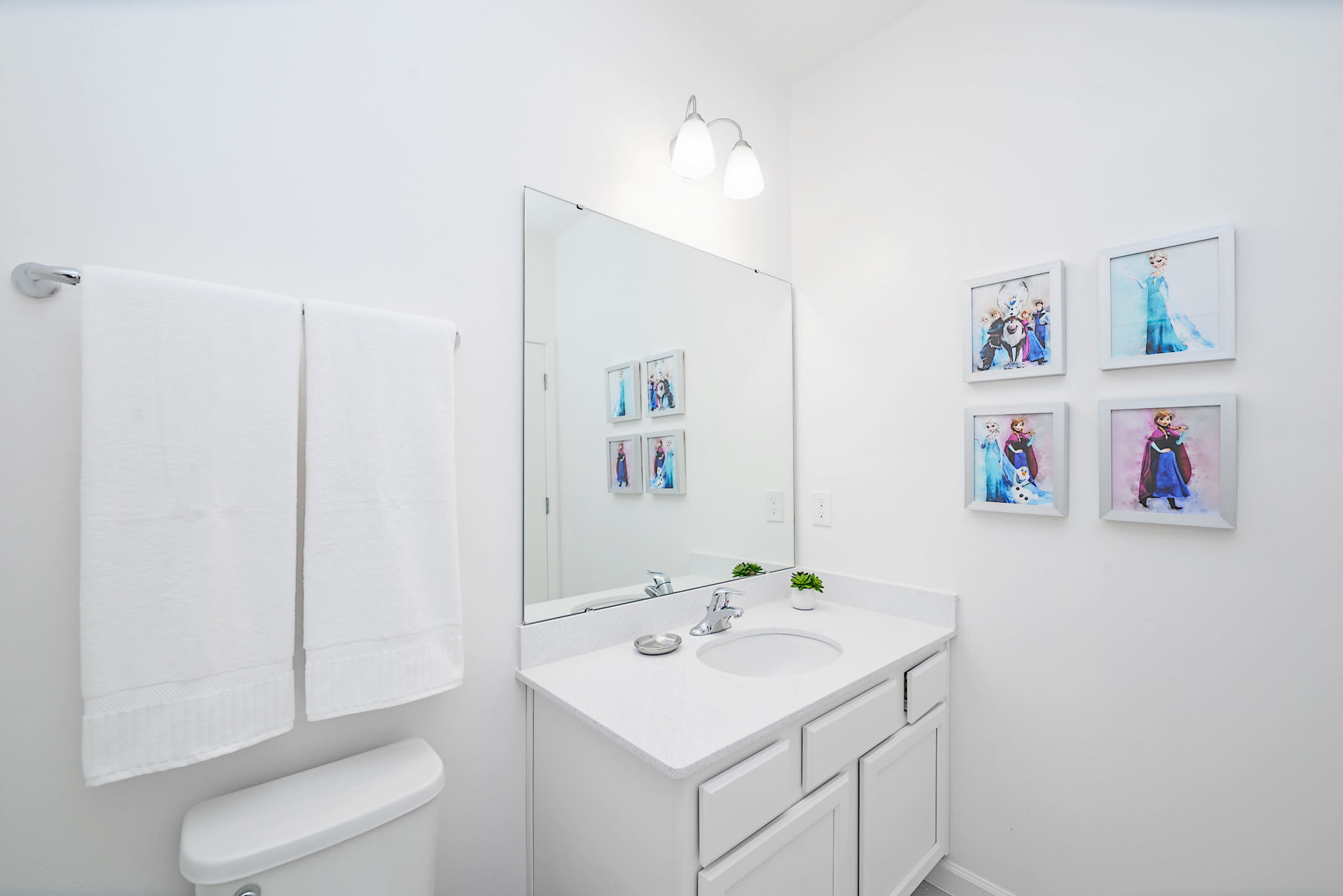 A pristine white bathroom equipped with a single vanity, a large mirror, a toilet, and framed Frozen character art on the wall. The room is functional and includes fresh white towels.