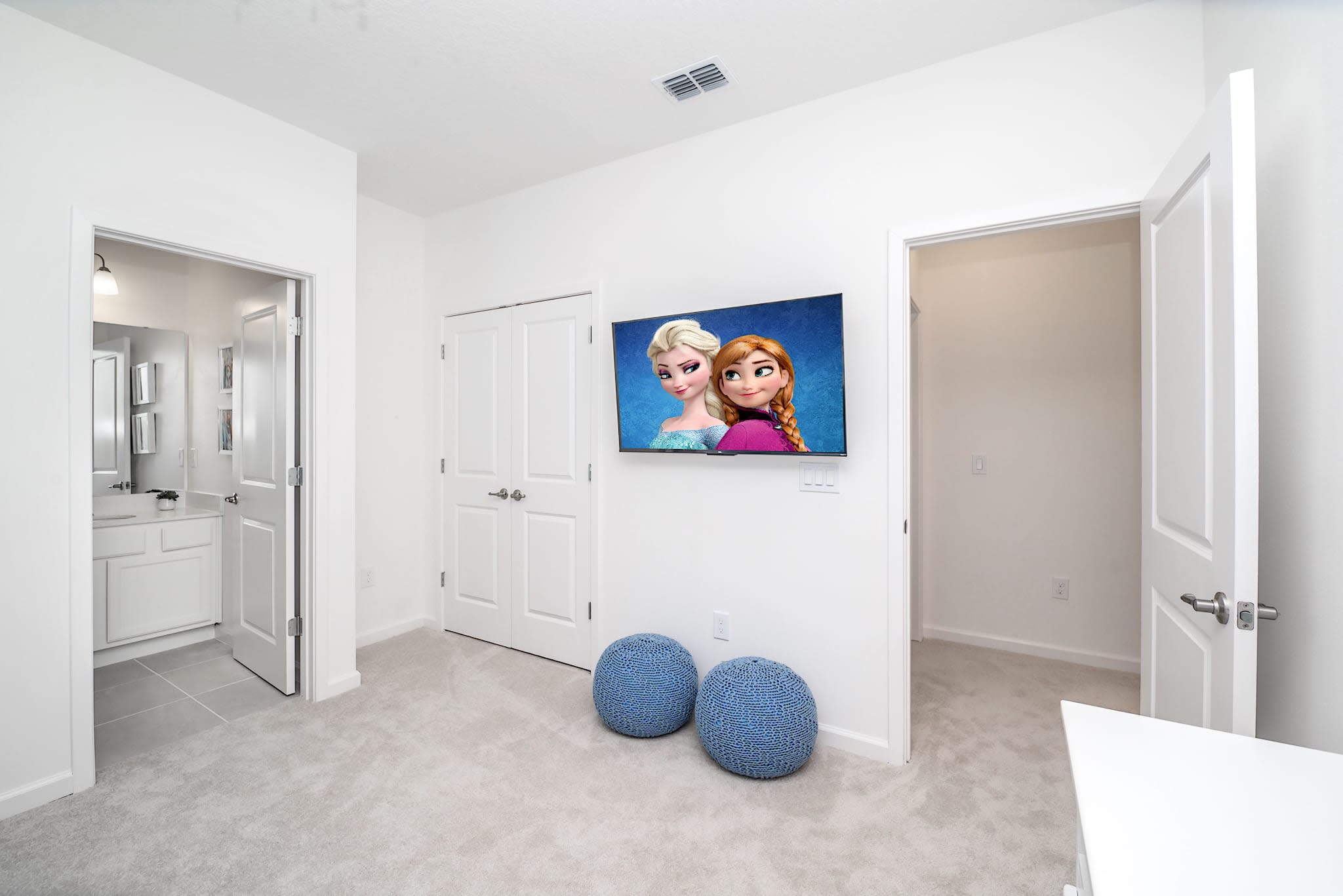 This view of the Frozen-themed bedroom highlights a wall-mounted TV displaying Frozen characters and two blue knitted poufs on the carpeted floor. Doorways lead to a closet and an en-suite bathroom.