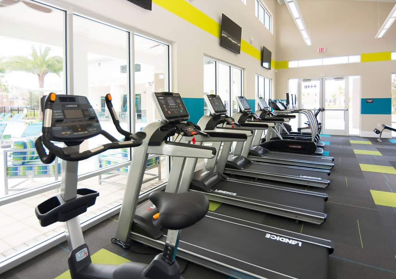 Close-up view of the cardio section in a modern fitness center, showing multiple treadmills and an exercise bike lined up against large windows. The floor features dark gray carpet tiles with vibrant lime green and blue accents.