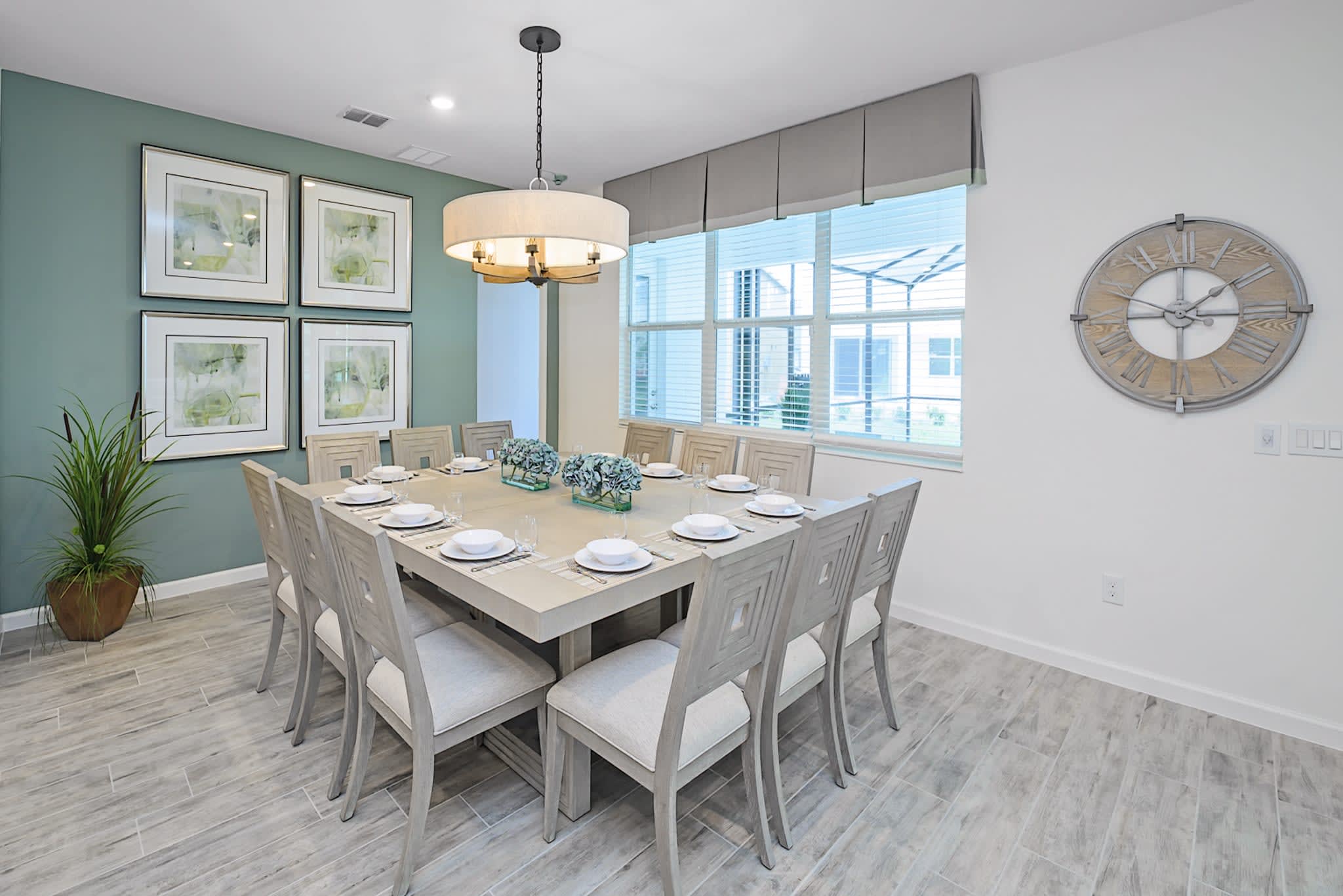 Another view of the dining area, highlighting the large square table with twelve chairs and a modern chandelier. A window with a valance provides natural light, and a decorative wall clock is visible on an adjacent wall.