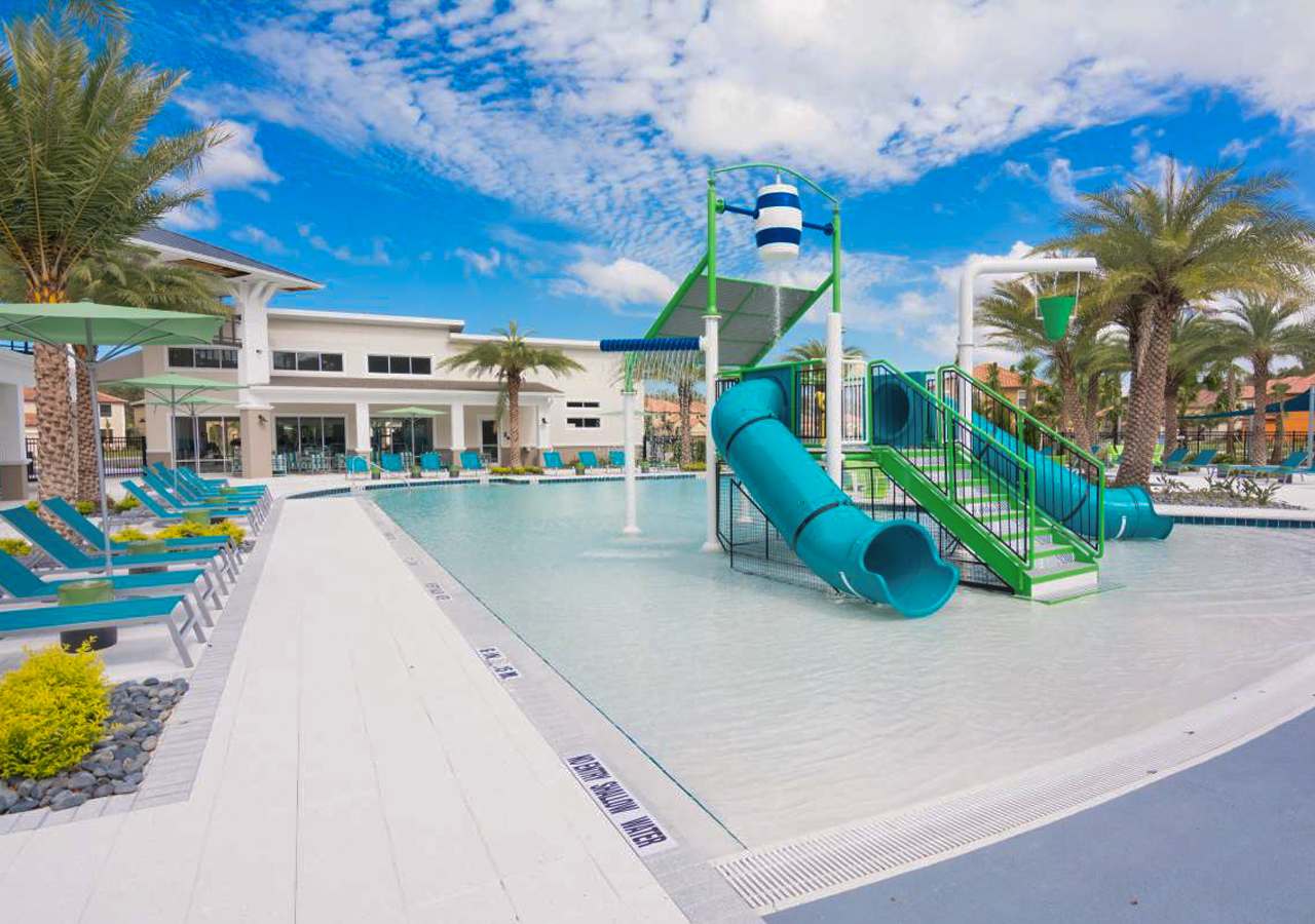 Expansive communal pool area with a shallow entry, a vibrant water play structure featuring slides and water features, surrounded by lounge chairs and palm trees under a blue sky.
