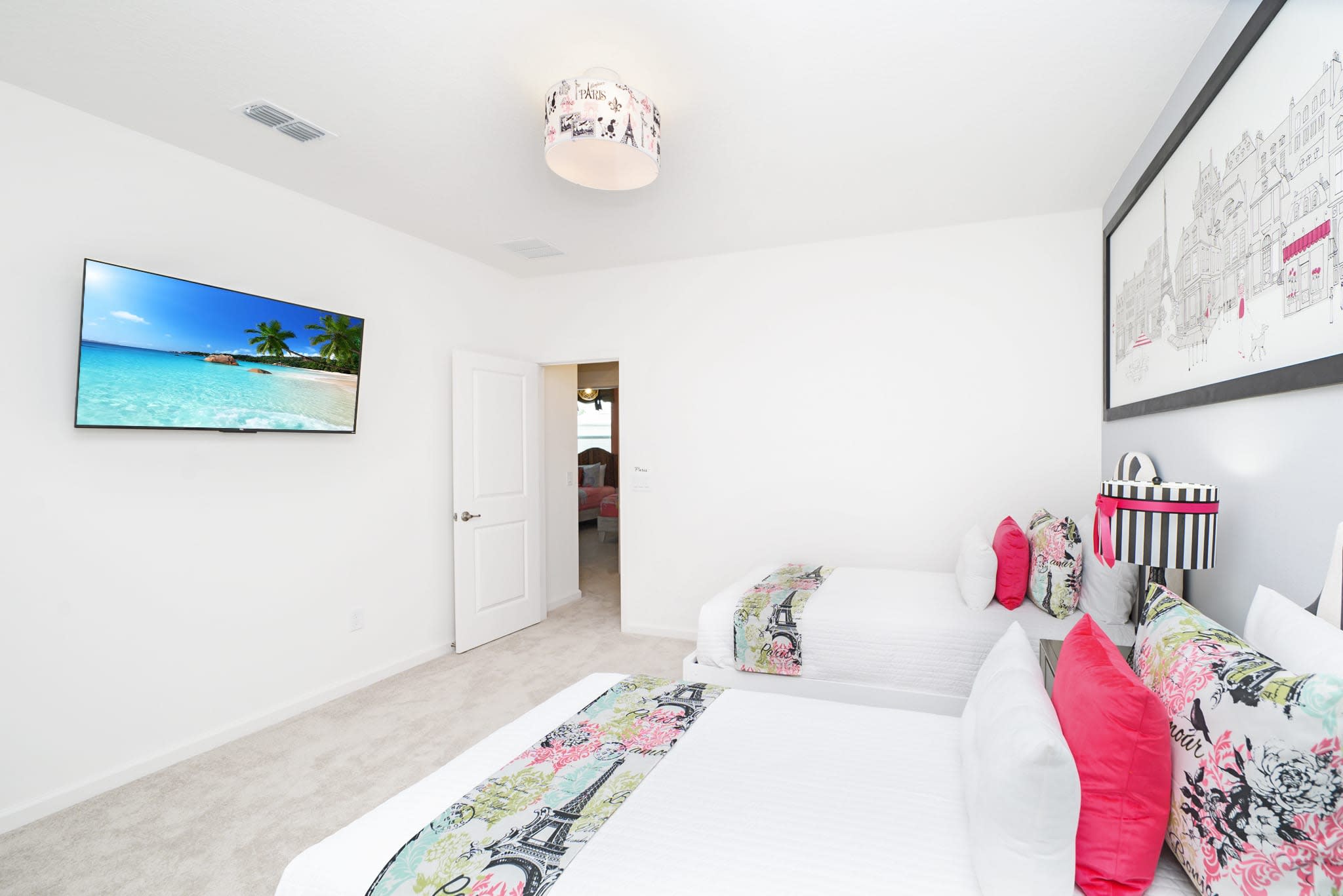 Another perspective of the Paris-themed bedroom, showcasing two twin beds adorned with floral and Eiffel Tower patterned runners. A wall-mounted TV displays a vibrant beach scene, and a doorway leads to an adjacent room.