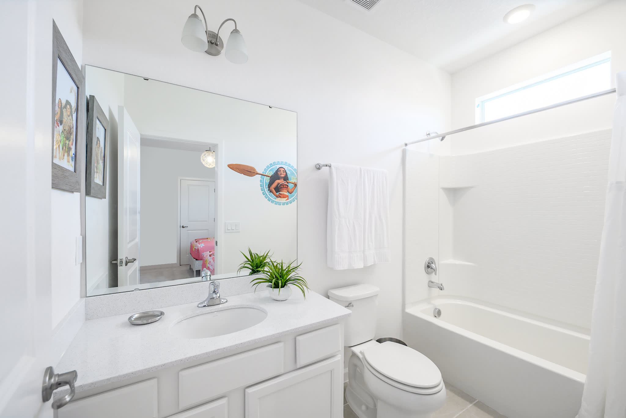 A clean white bathroom with a single vanity, a large mirror, a toilet, and a bathtub with a shower curtain. Two white towels hang on a bar, and a small window is above the tub.