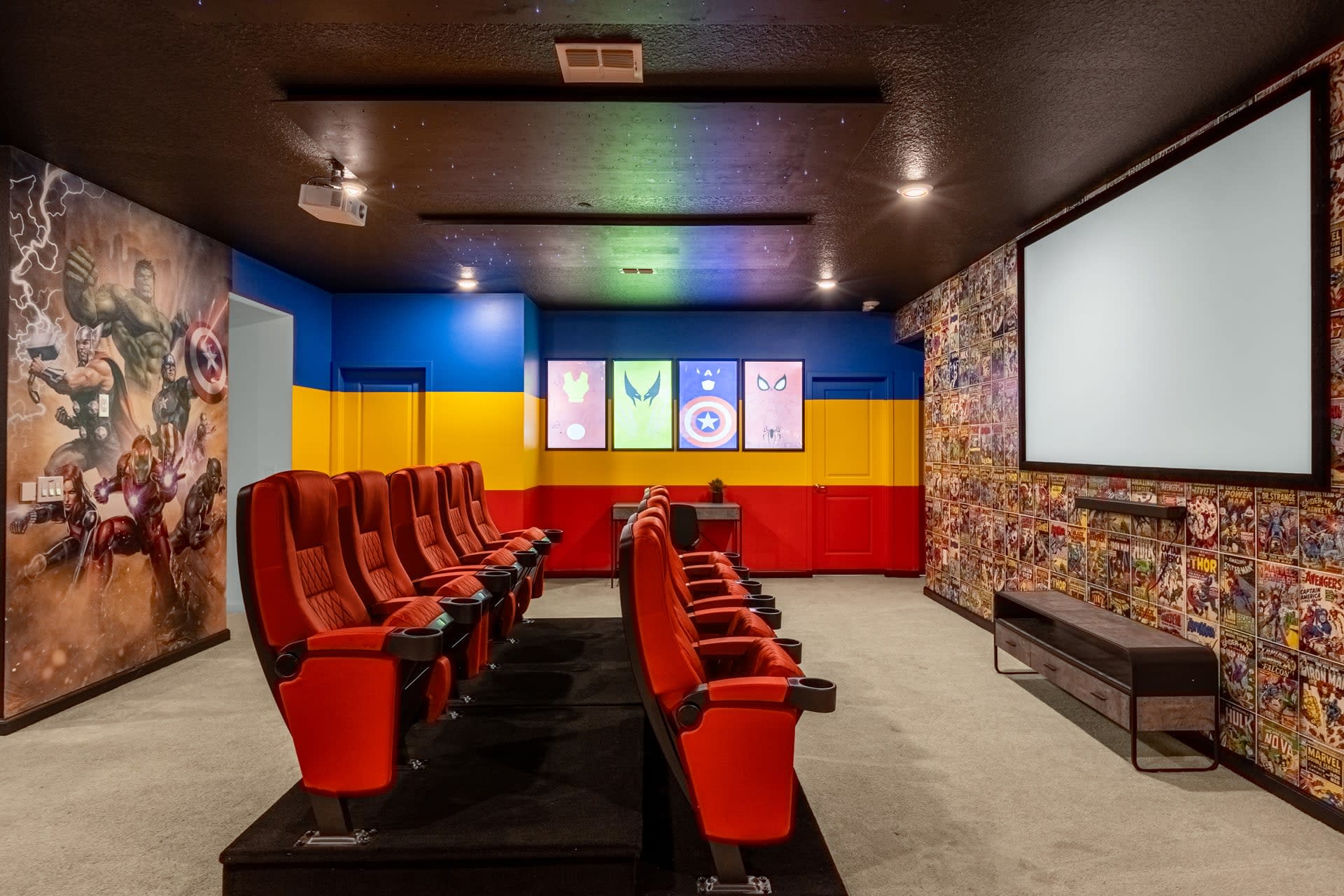 A dedicated home cinema room with two rows of plush red theater seats facing a large projection screen. The walls feature superhero murals and comic book wallpaper, with a dark ceiling and recessed lighting.