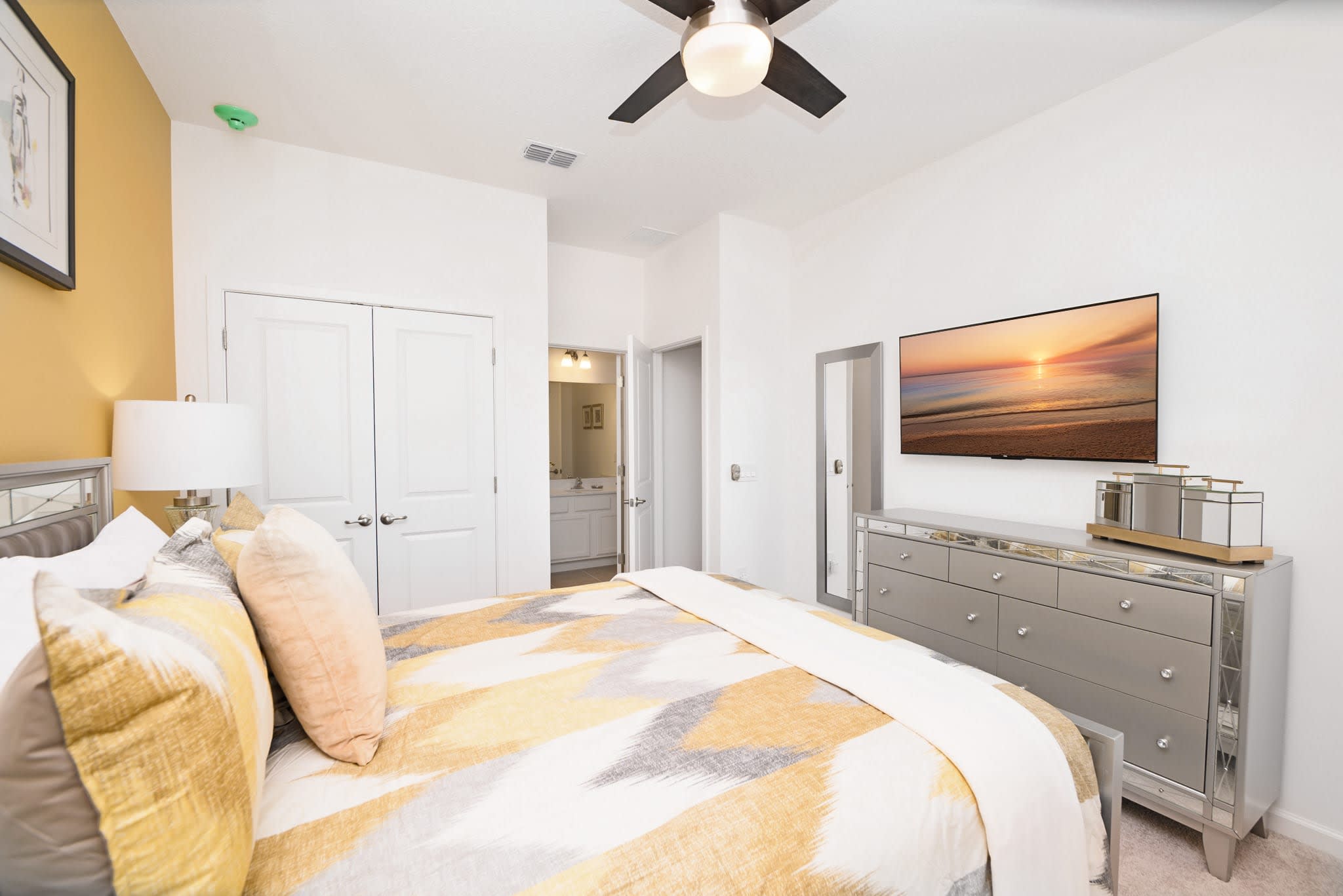A modern bedroom with a queen-sized bed featuring a geometric patterned duvet, mirrored nightstands, and a wall-mounted TV above a mirrored dresser. A full-length mirror is visible near the bathroom entrance.