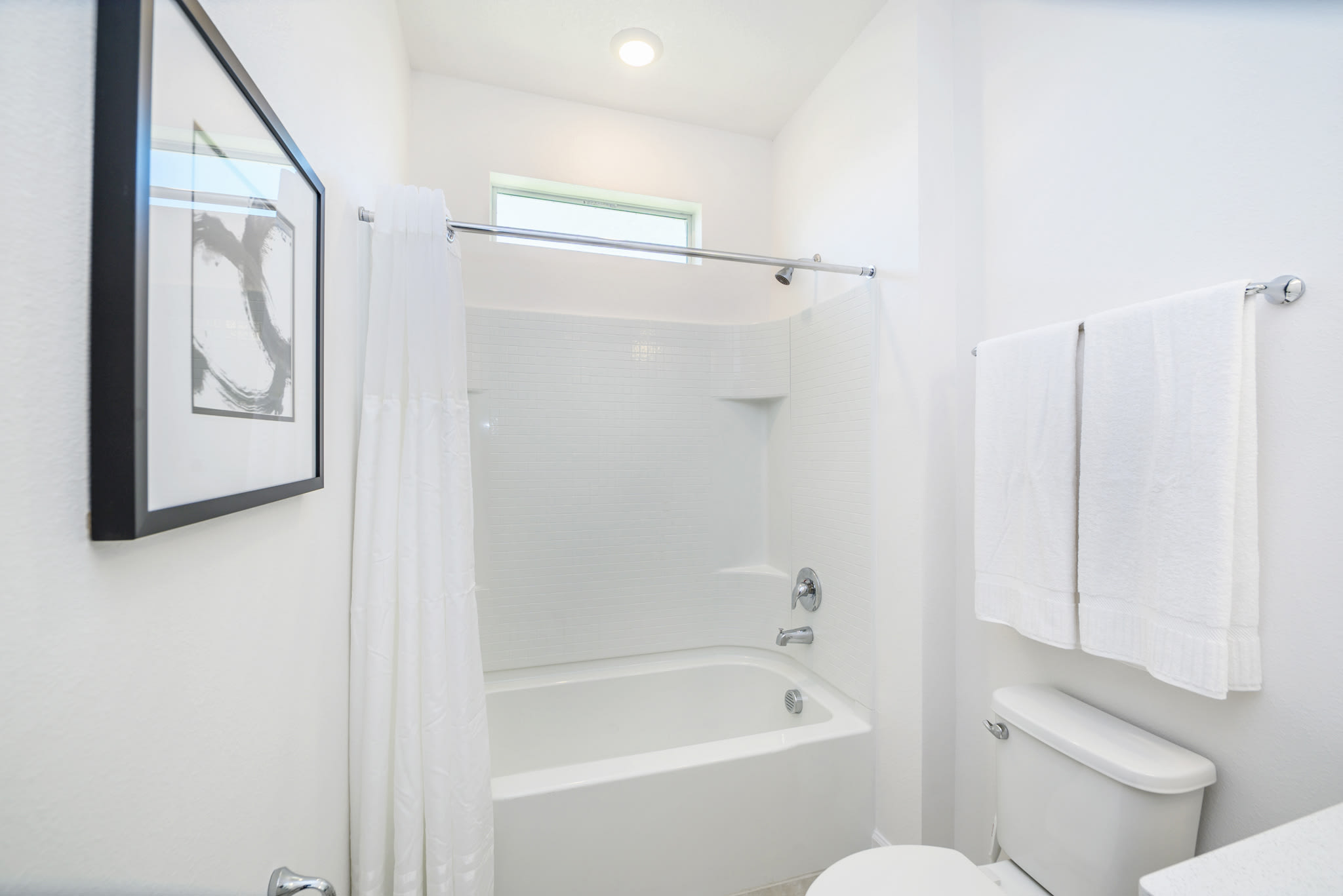 A bright, minimalist bathroom featuring a white bathtub with a shower curtain, a toilet, and a small window above the tub. The walls are white, and a framed abstract artwork hangs on one wall.