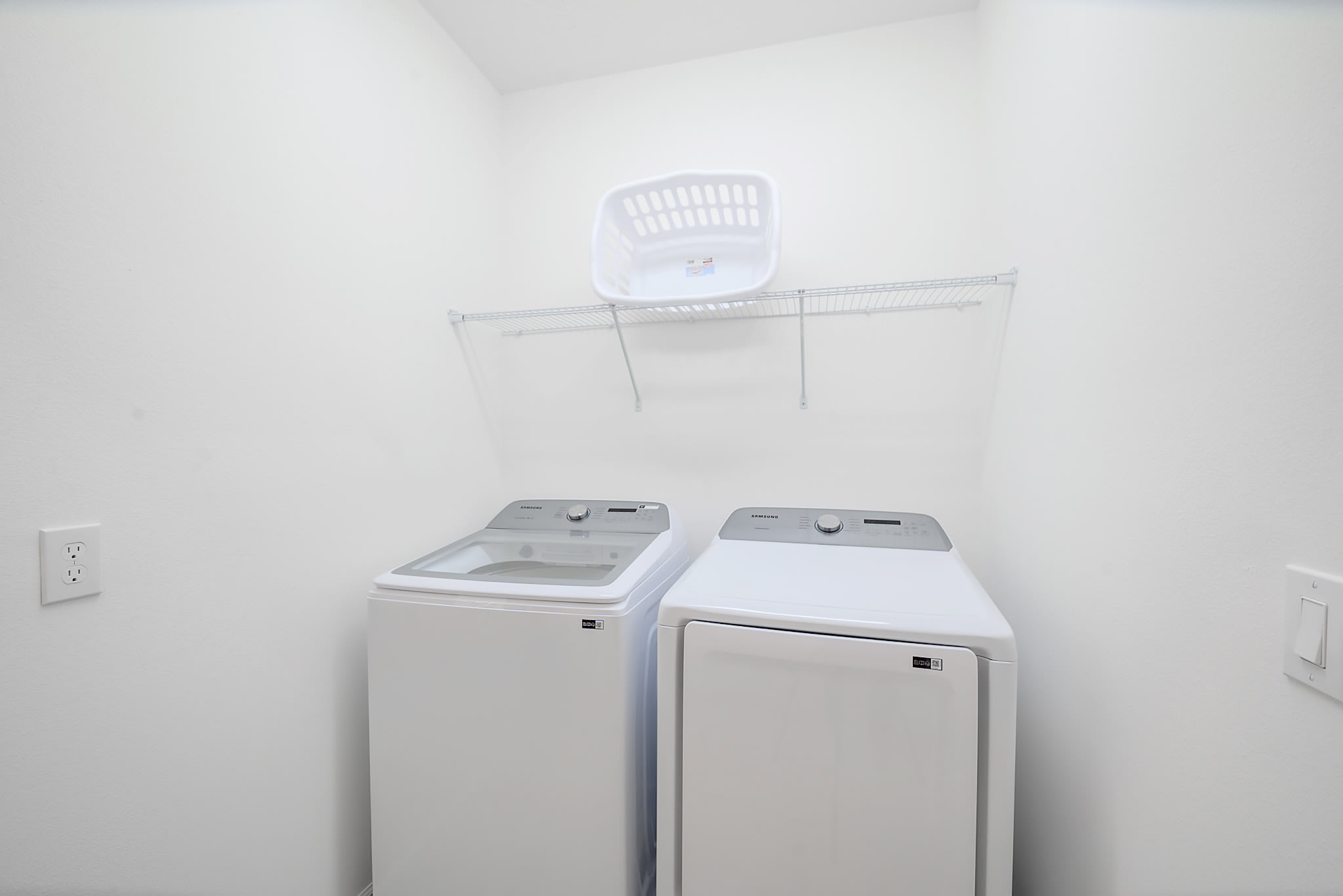 Functional laundry room with white walls, a top-loading washing machine, a front-loading dryer, and a wire shelf with a laundry basket above. The room is compact and well-lit.