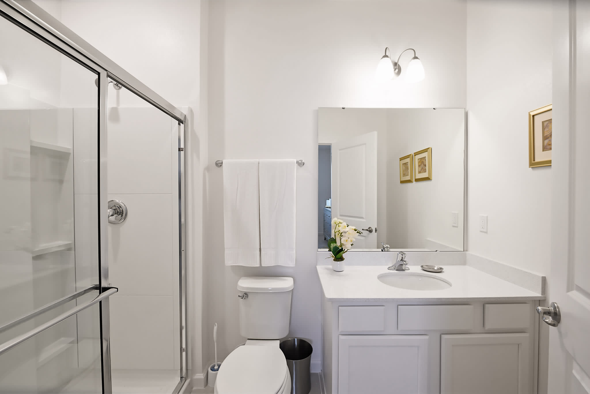 A bright, modern bathroom featuring a glass-enclosed shower, a white toilet, and a vanity with a white countertop and sink. White towels are neatly folded on a rack above the toilet.
