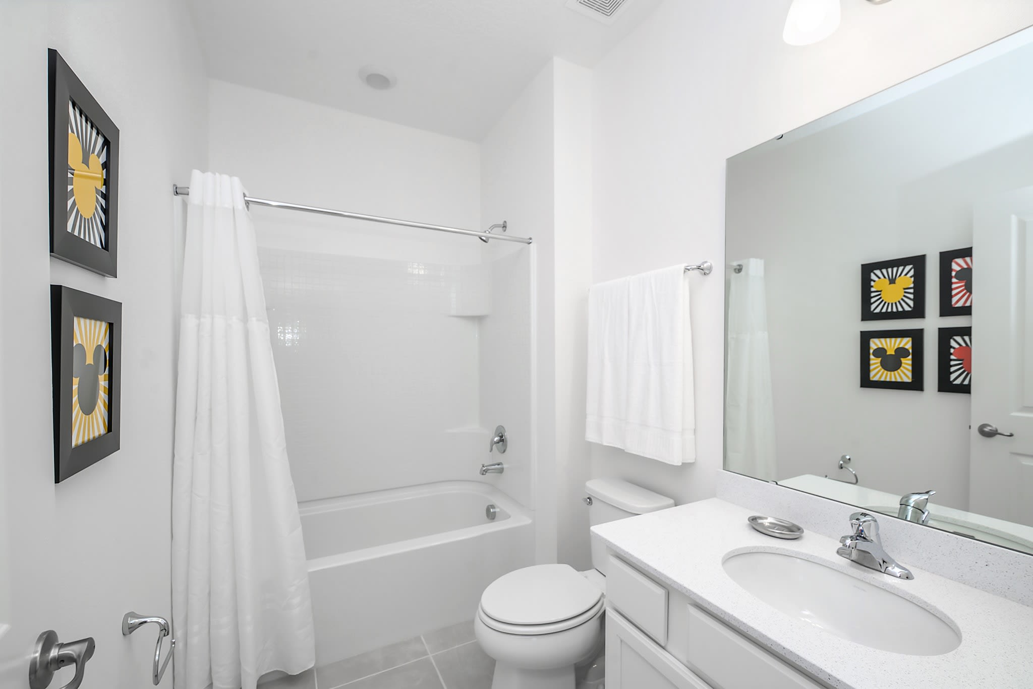 Bright white bathroom featuring a shower-tub combo with a white curtain, a toilet, a vanity with a large mirror, and framed Mickey Mouse silhouette artwork on the wall. The floor is light grey tile.
