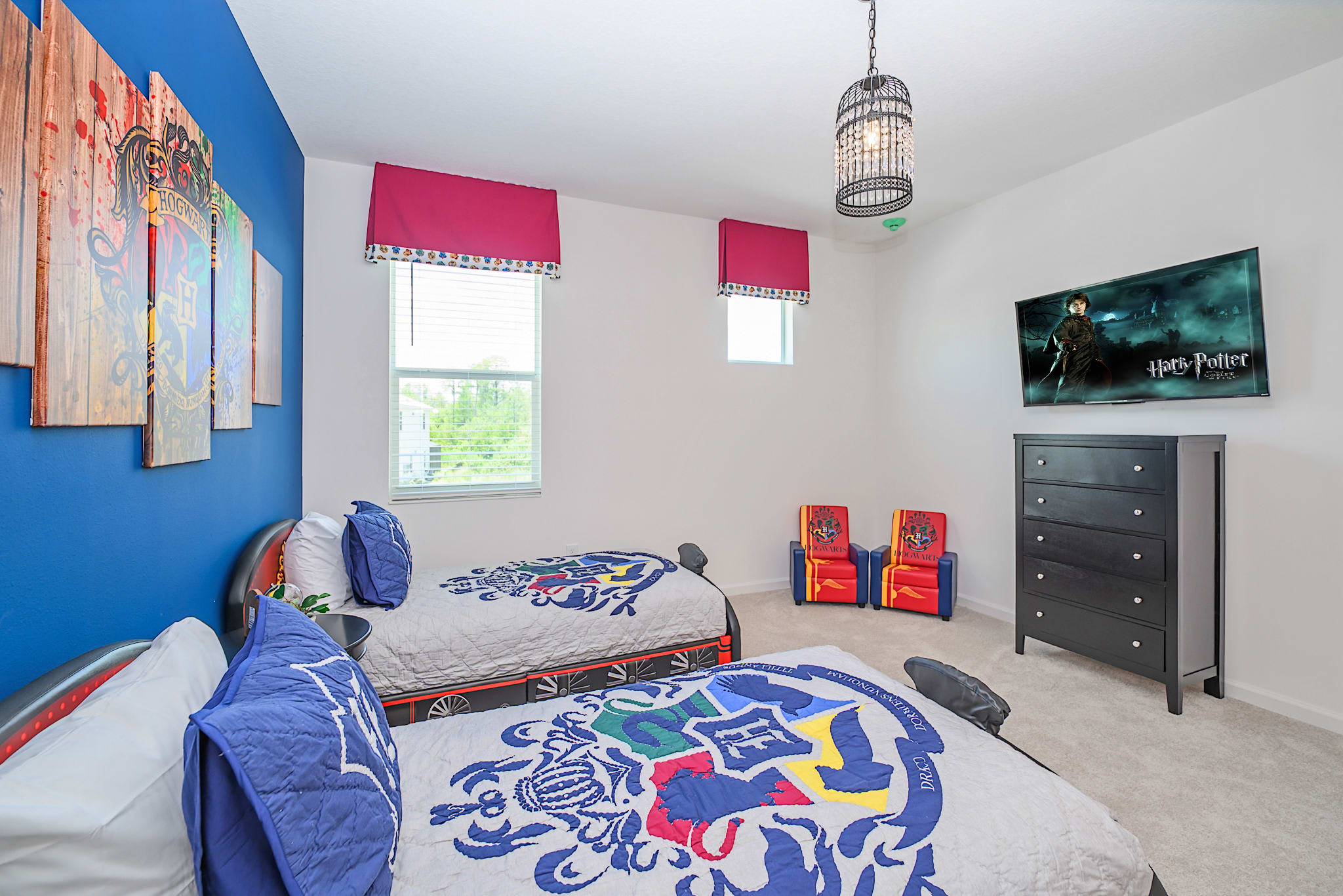 A Harry Potter themed bedroom featuring two twin beds with themed bedding, a vibrant blue accent wall, and a wall-mounted TV displaying Harry Potter content. A black dresser and a unique birdcage light fixture complete the magical ambiance.