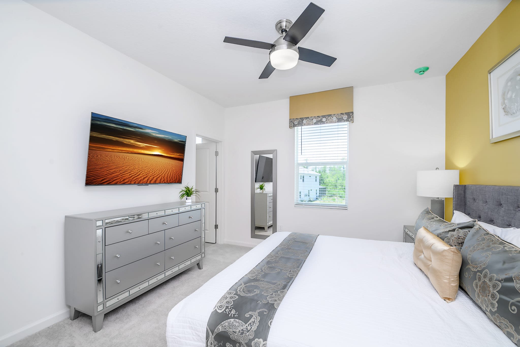 View of a bright bedroom with a wall-mounted TV above a mirrored dresser, facing a king-sized bed with a gray upholstered headboard. A window with blinds offers natural light, and a ceiling fan is visible.
