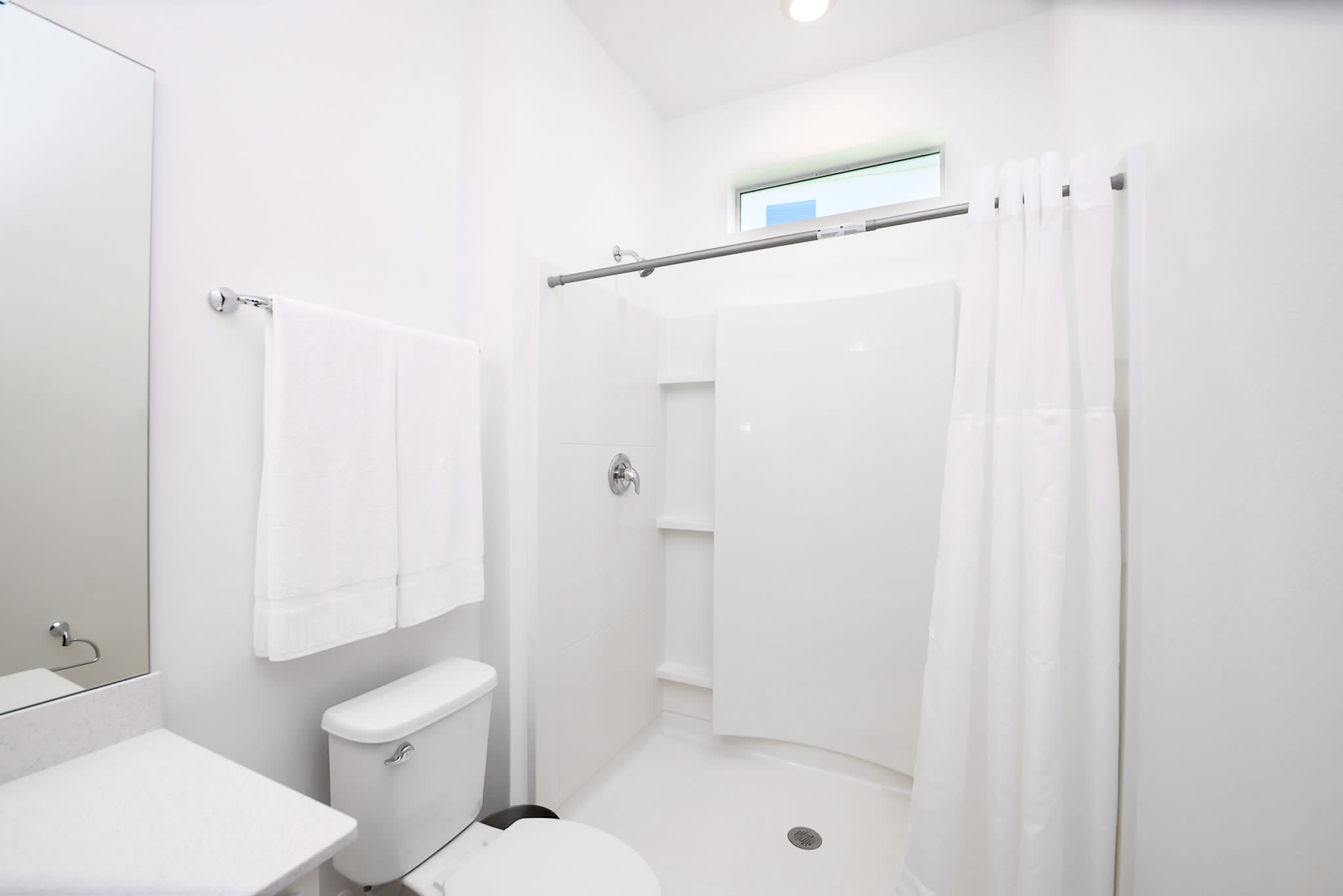 A clean, modern bathroom with a white stand-up shower stall, a toilet, and a single vanity with a mirror. A small horizontal window is positioned high on the wall above the shower.