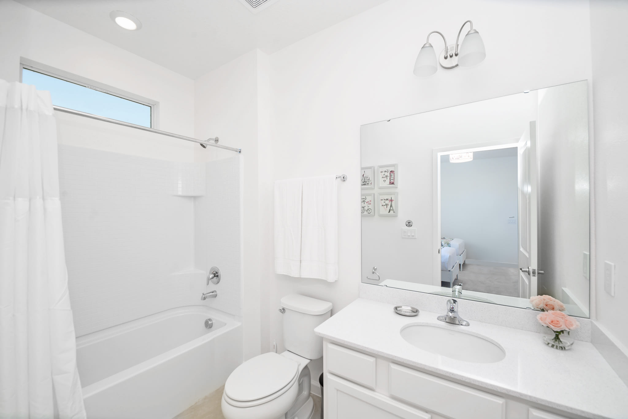 A bright white bathroom featuring a single vanity with a large mirror, a toilet, and a bathtub with a shower. The room is decorated with framed Paris-themed art and includes a small horizontal window for natural light.