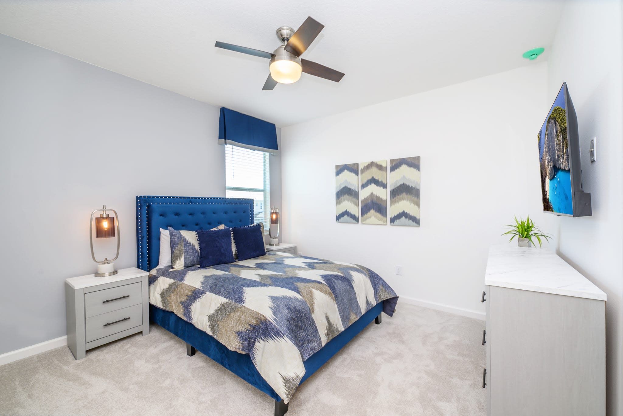 A stylish bedroom featuring a queen-sized bed with a blue upholstered headboard and a patterned blue and white comforter. A window with a blue valance provides natural light, and a ceiling fan is overhead.