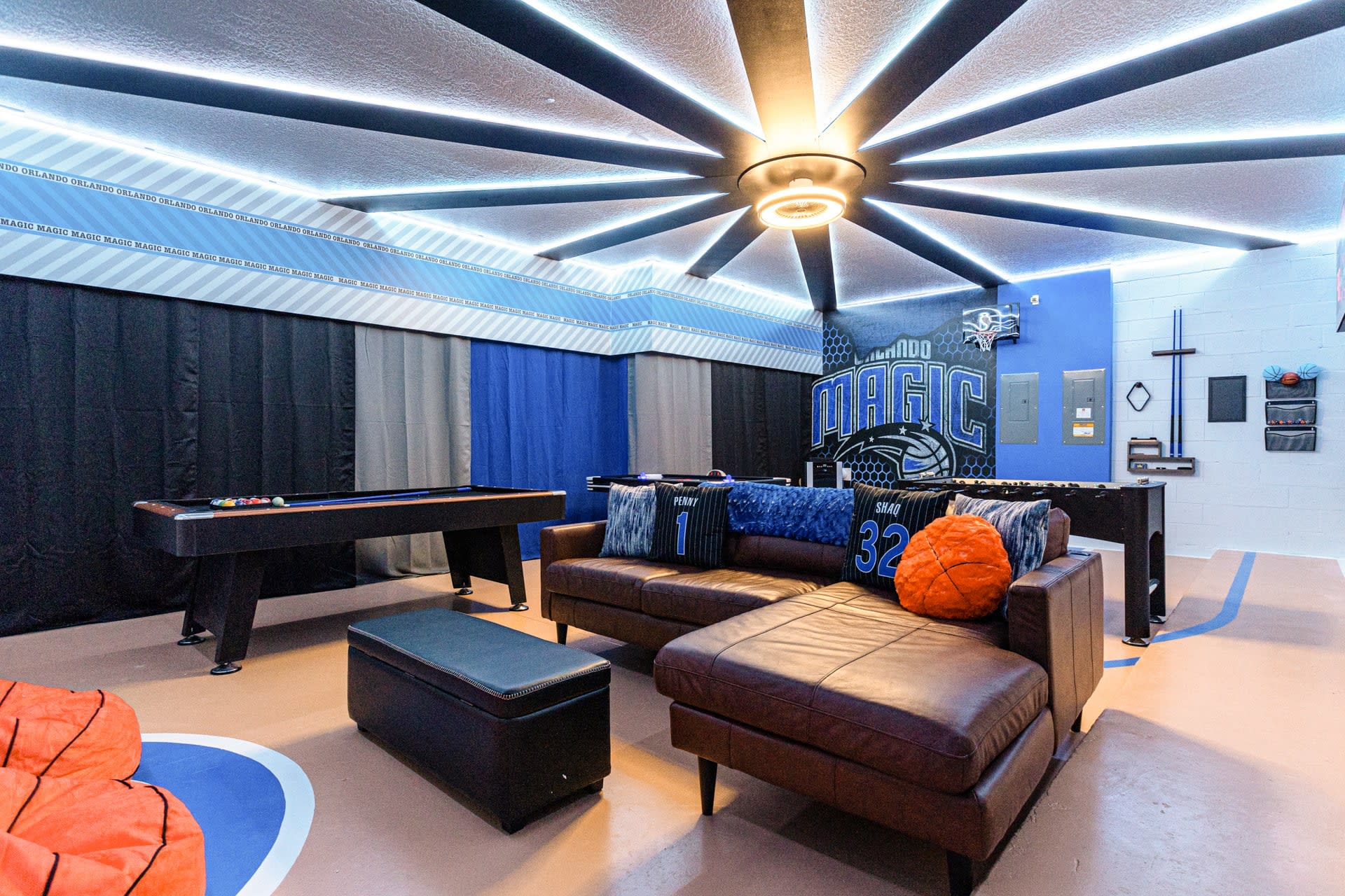 A spacious game room featuring a pool table, a comfortable brown leather sectional sofa with themed pillows, and a foosball table. The room has a basketball court-themed floor and a ceiling with radial LED lighting.