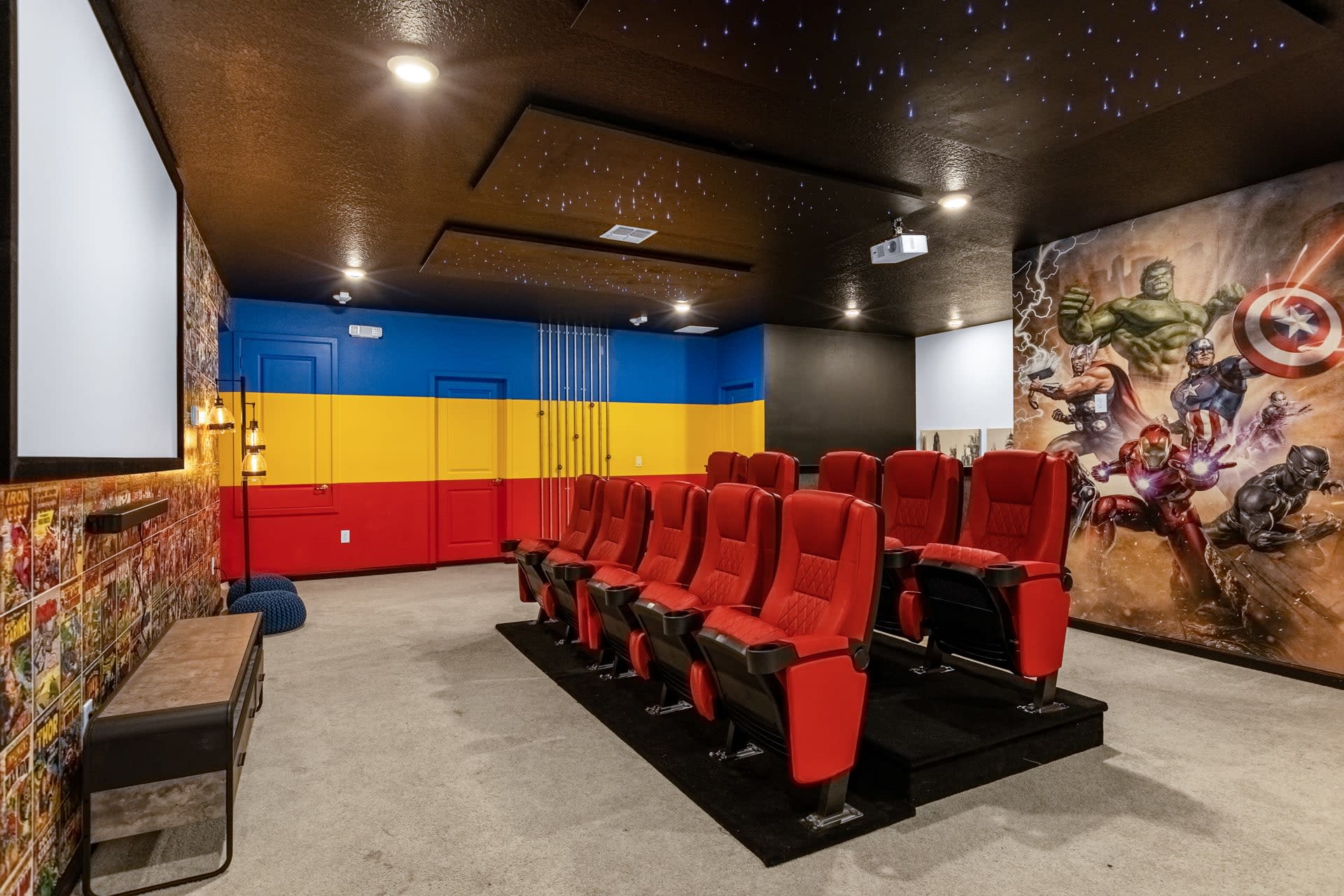 The media room features multiple rows of red cinema-style seating facing a large projection screen. One wall is adorned with a vibrant Marvel superhero mural, while the ceiling is dark with twinkling star lights.