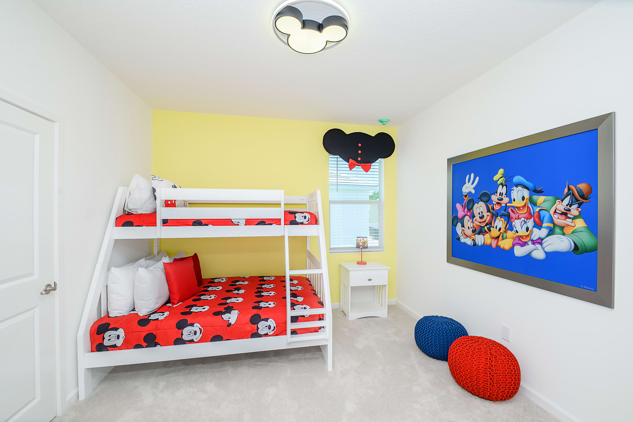 Kids' bedroom with white bunk beds featuring red Mickey Mouse bedding, a yellow accent wall, and a large framed Mickey Mouse and friends artwork. The room has light grey carpet and a white nightstand.