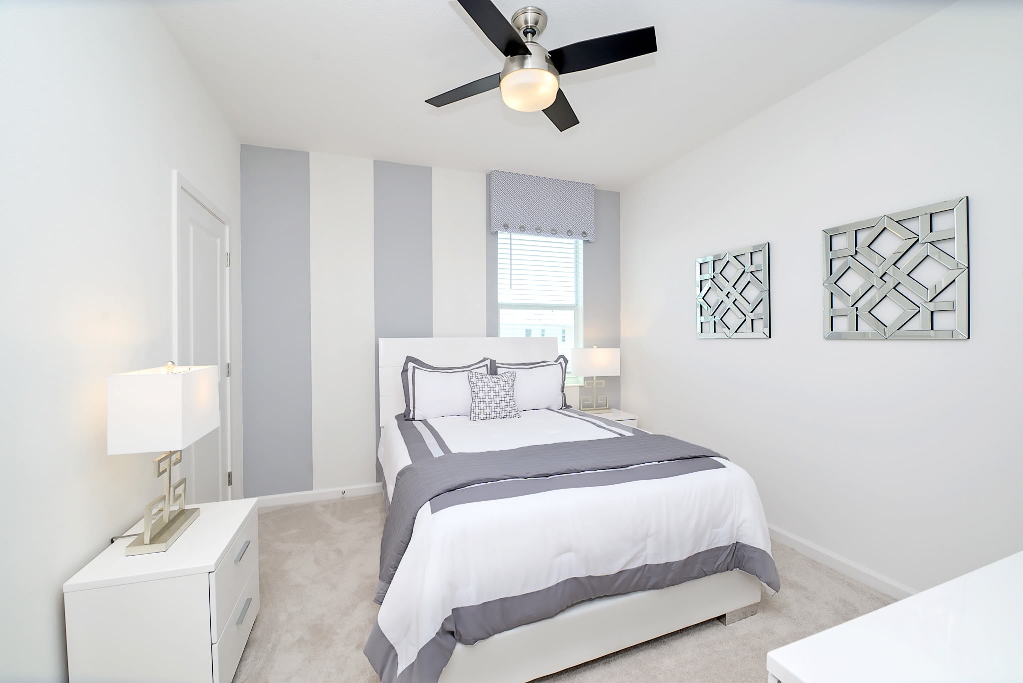 A modern bedroom featuring a white bed with gray and white bedding, flanked by white nightstands with lamps. A striking accent wall with wide gray and white vertical stripes is behind the bed, and a ceiling fan with black blades hangs overhead.