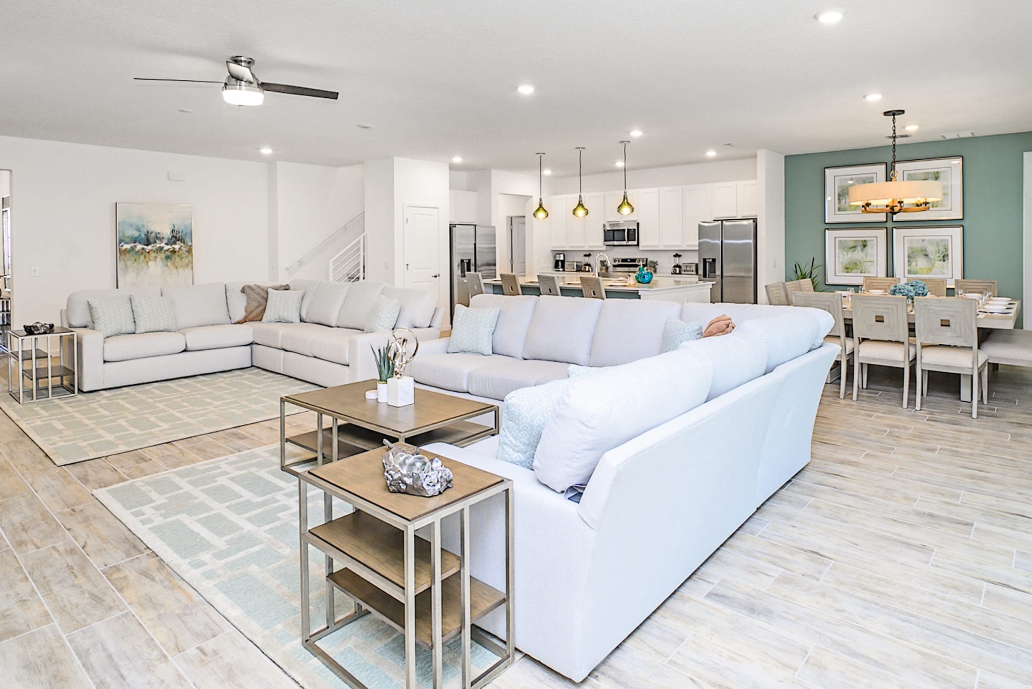 A spacious open-concept living area with two large L-shaped white sofas and light-colored tile flooring. The room flows into a modern kitchen with white cabinetry and a dining area with a light green accent wall.