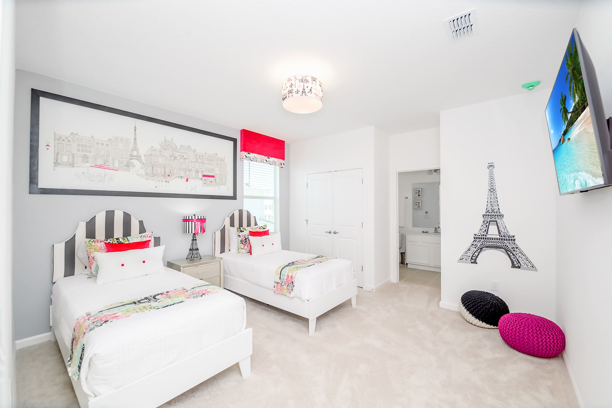 A Paris-themed bedroom with two twin beds featuring distinctive striped headboards, a large framed cityscape artwork, and an Eiffel Tower wall decal. The room is accented with red window treatments and includes a wall-mounted TV.