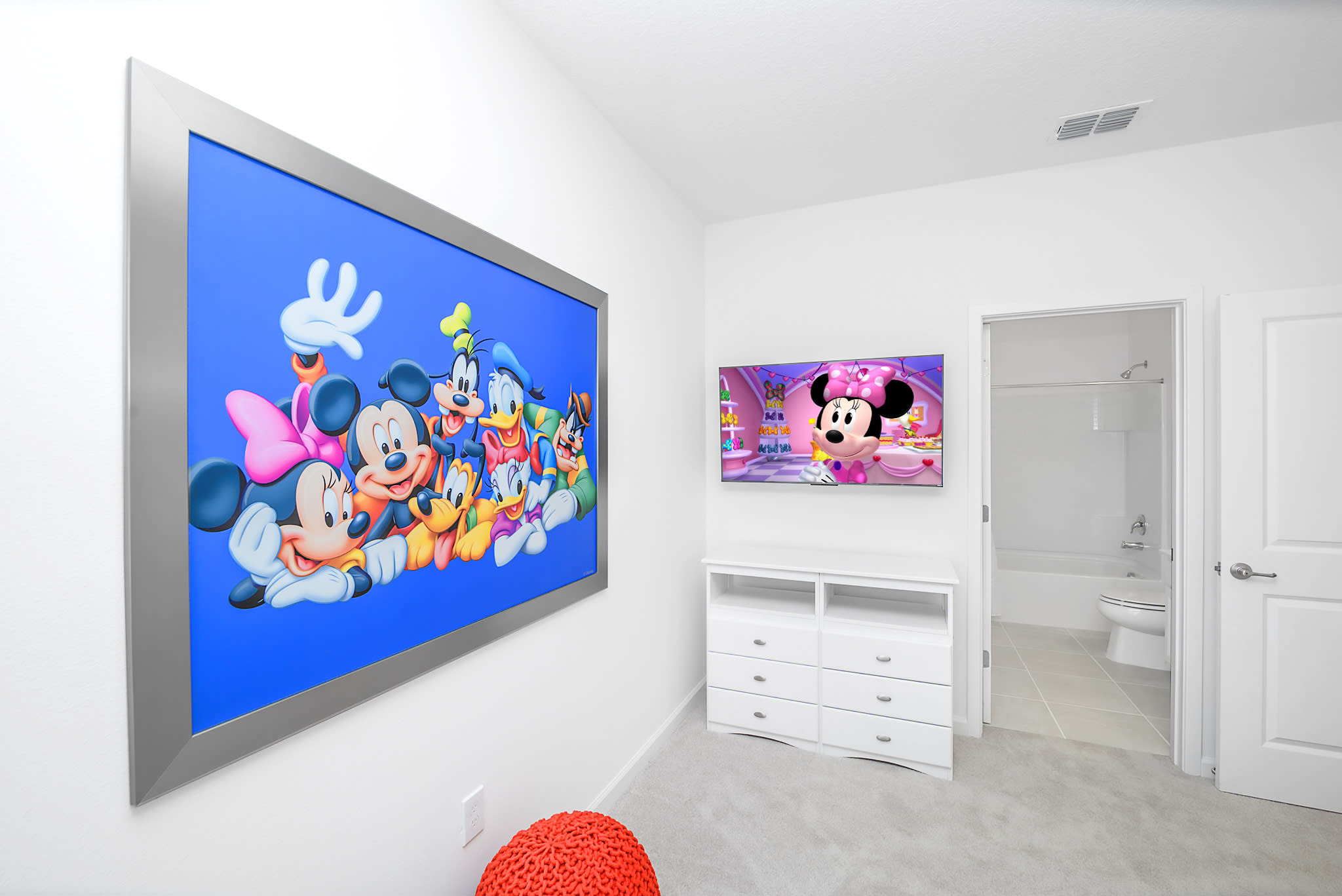 Continuation of the Mickey Mouse themed kids' bedroom, showing a wall-mounted TV displaying Minnie Mouse, a white dresser, and a large framed Mickey Mouse and friends artwork. Two colorful pouf ottomans sit on the carpet.
