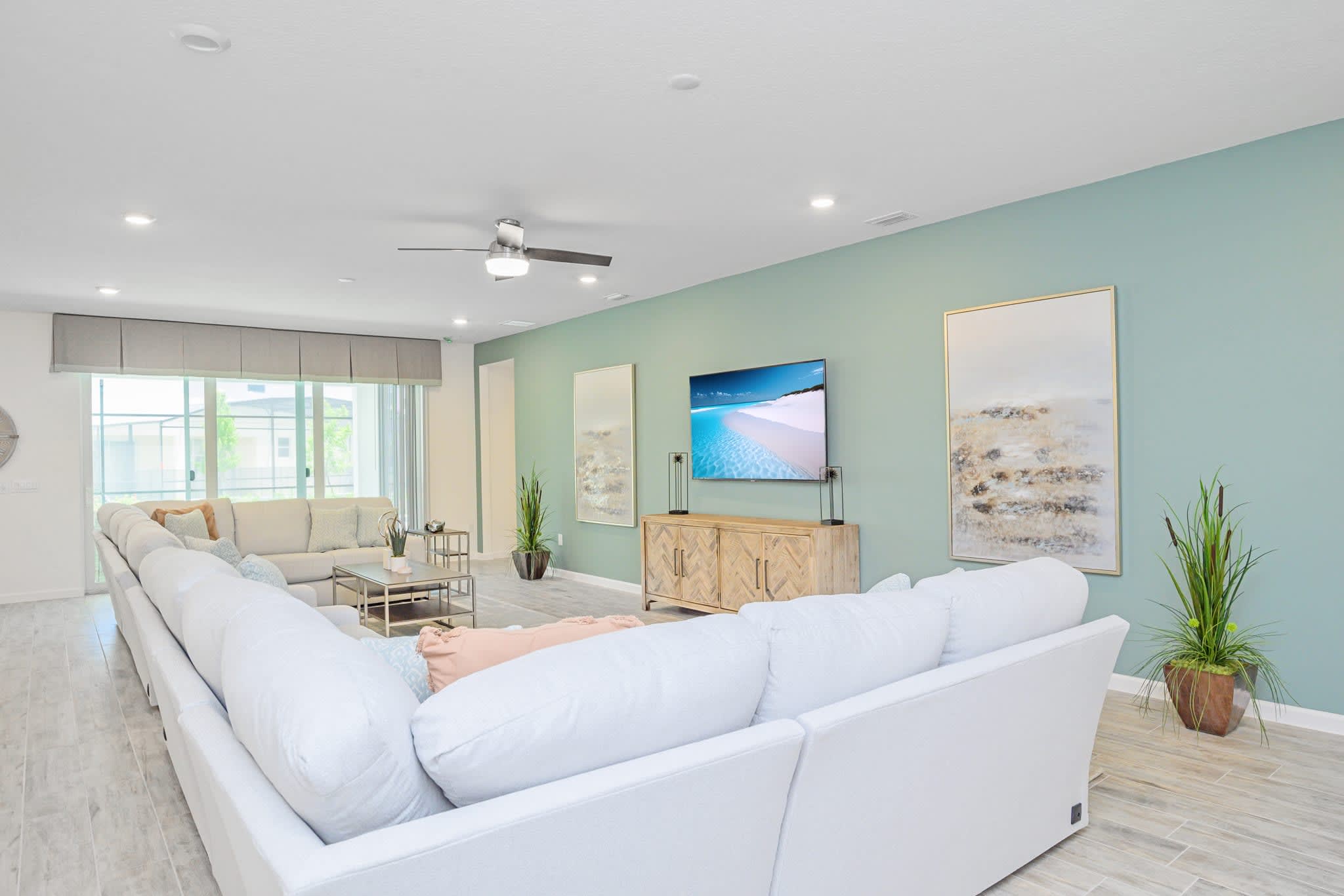 The living room features a large wall-mounted TV above a rustic wooden console, set against a light teal accent wall. A spacious white sectional sofa provides ample seating, with light wood-look tile floors and large windows.