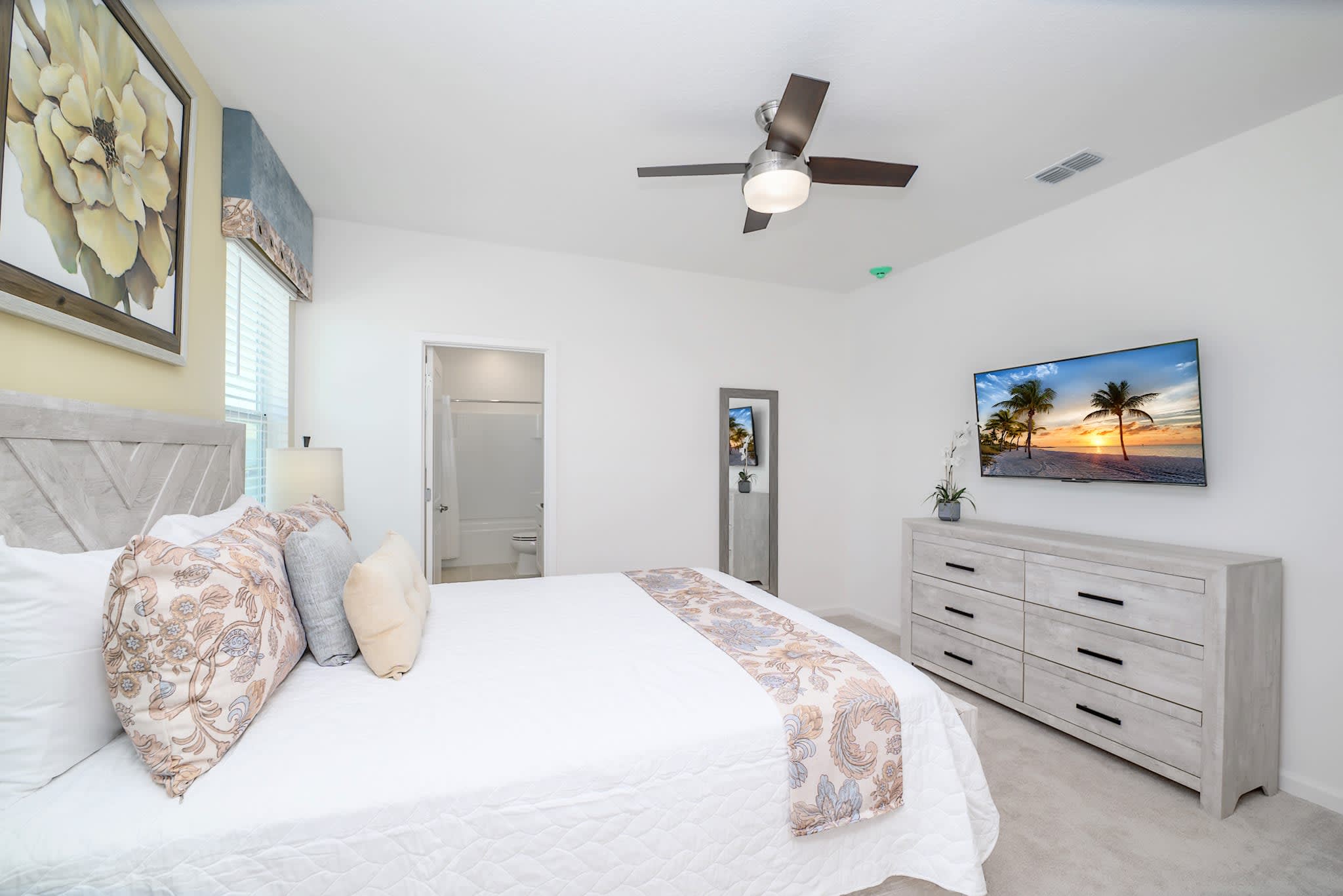 View of a bright bedroom with a queen-sized bed, a wall-mounted TV above a light wood dresser, and a ceiling fan. A doorway leads to an en-suite bathroom, and a full-length mirror is visible.