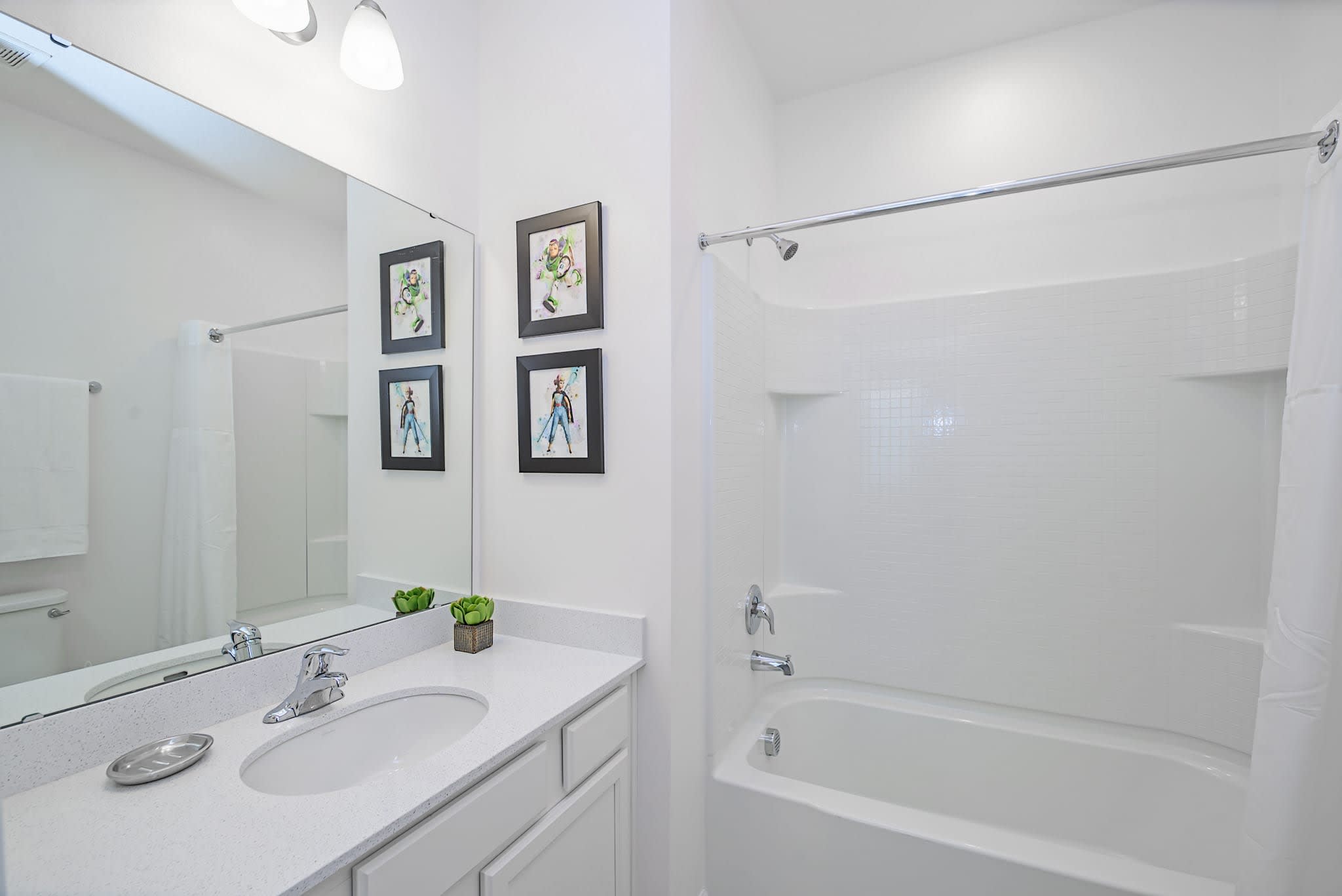 A clean, white bathroom with a single vanity, a large mirror, a toilet, and a bathtub with a shower. The room is decorated with framed art featuring colorful characters and a small green plant on the counter.