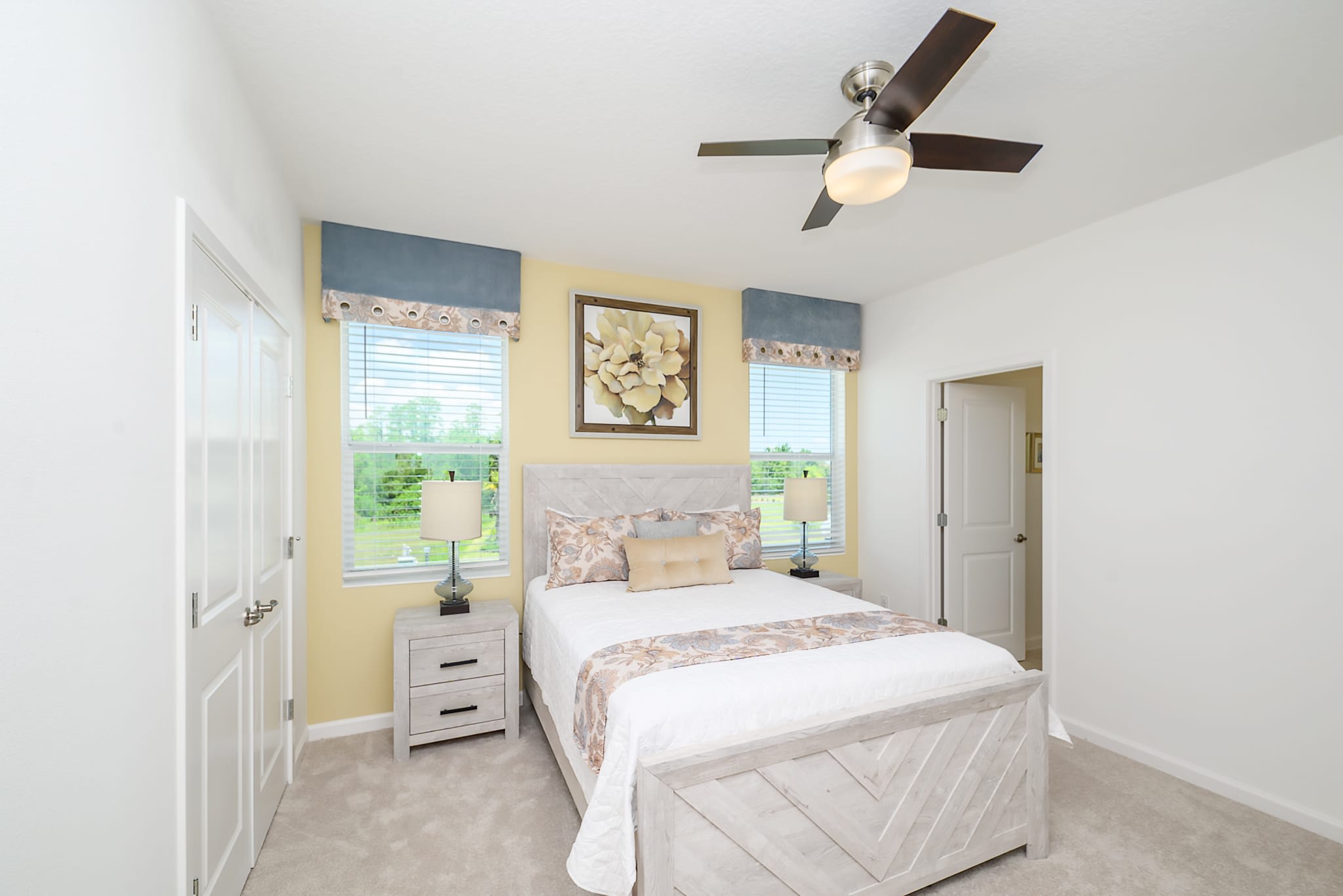 A cozy bedroom featuring a queen-sized bed with a light wood frame and a decorative throw. Two windows with blinds flank a large framed floral artwork on a yellow accent wall, and a ceiling fan is mounted overhead.
