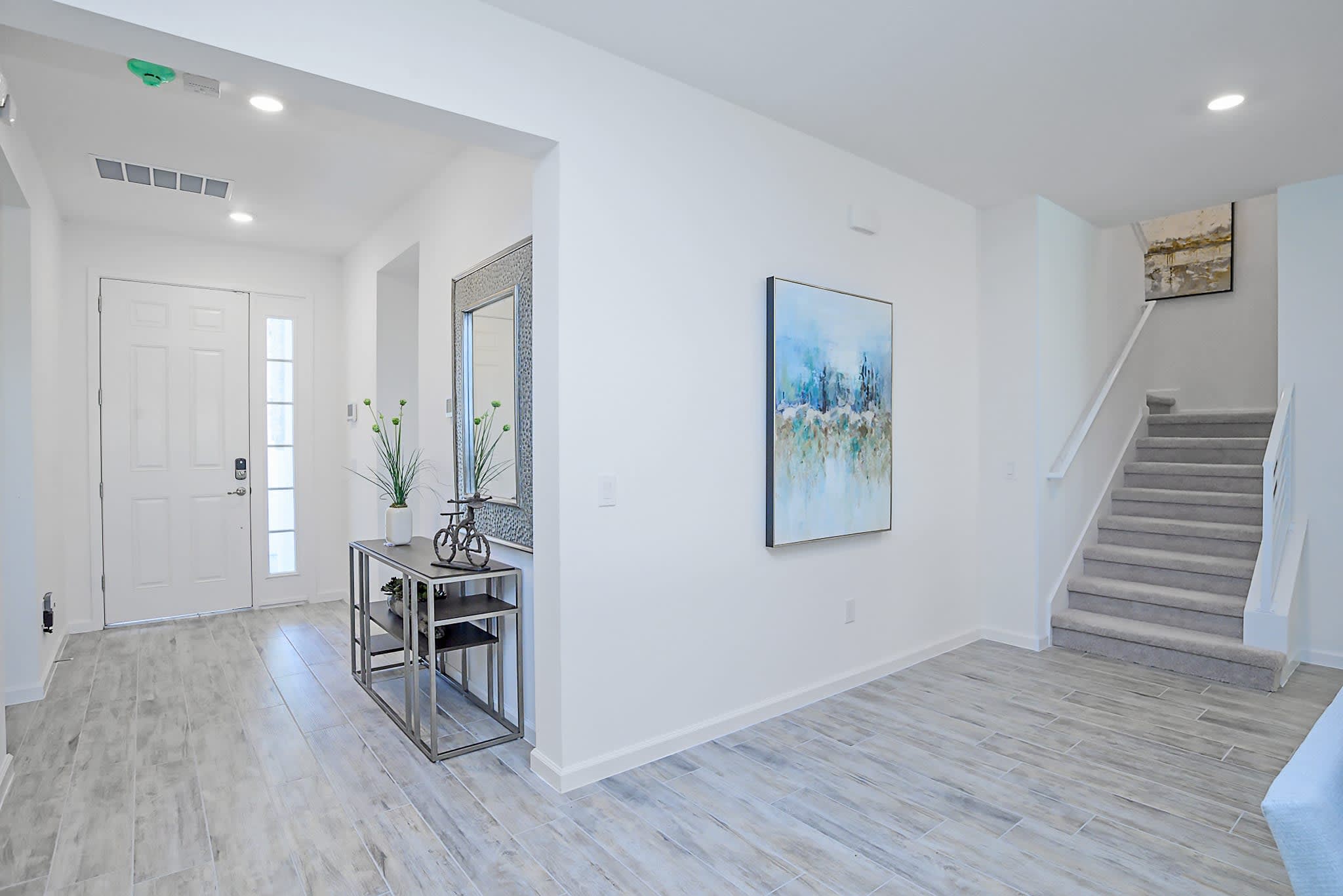 Bright entryway with light wood-look tile flooring, a white front door, a console table with a large decorative mirror, and a carpeted staircase leading upstairs. Modern artwork hangs on the adjacent wall.