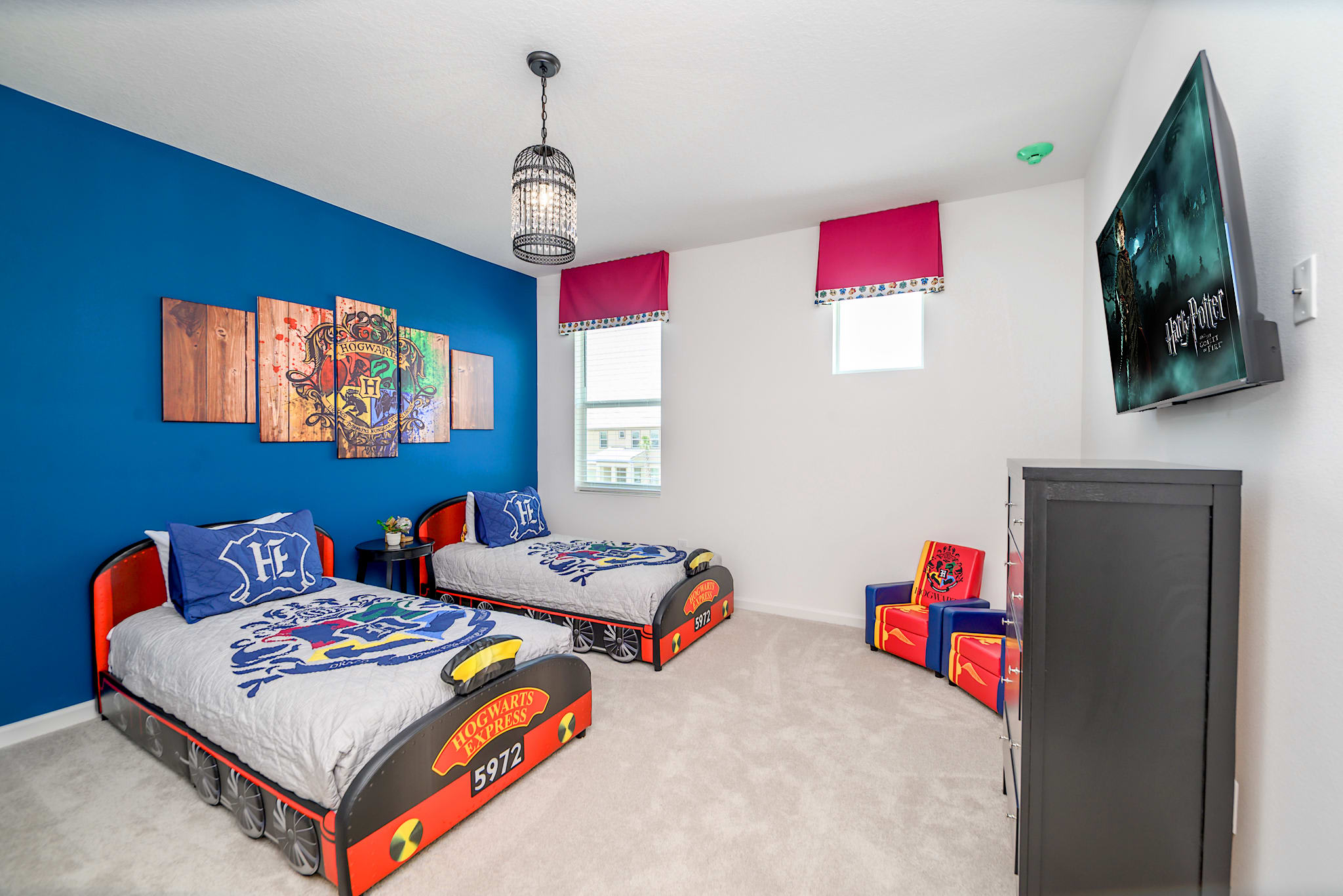 A Harry Potter-themed bedroom with two twin beds designed to look like train cars, featuring blue and red bedding. A large multi-panel Harry Potter wall art is on a vibrant blue accent wall, and a unique caged chandelier hangs from the ceiling.