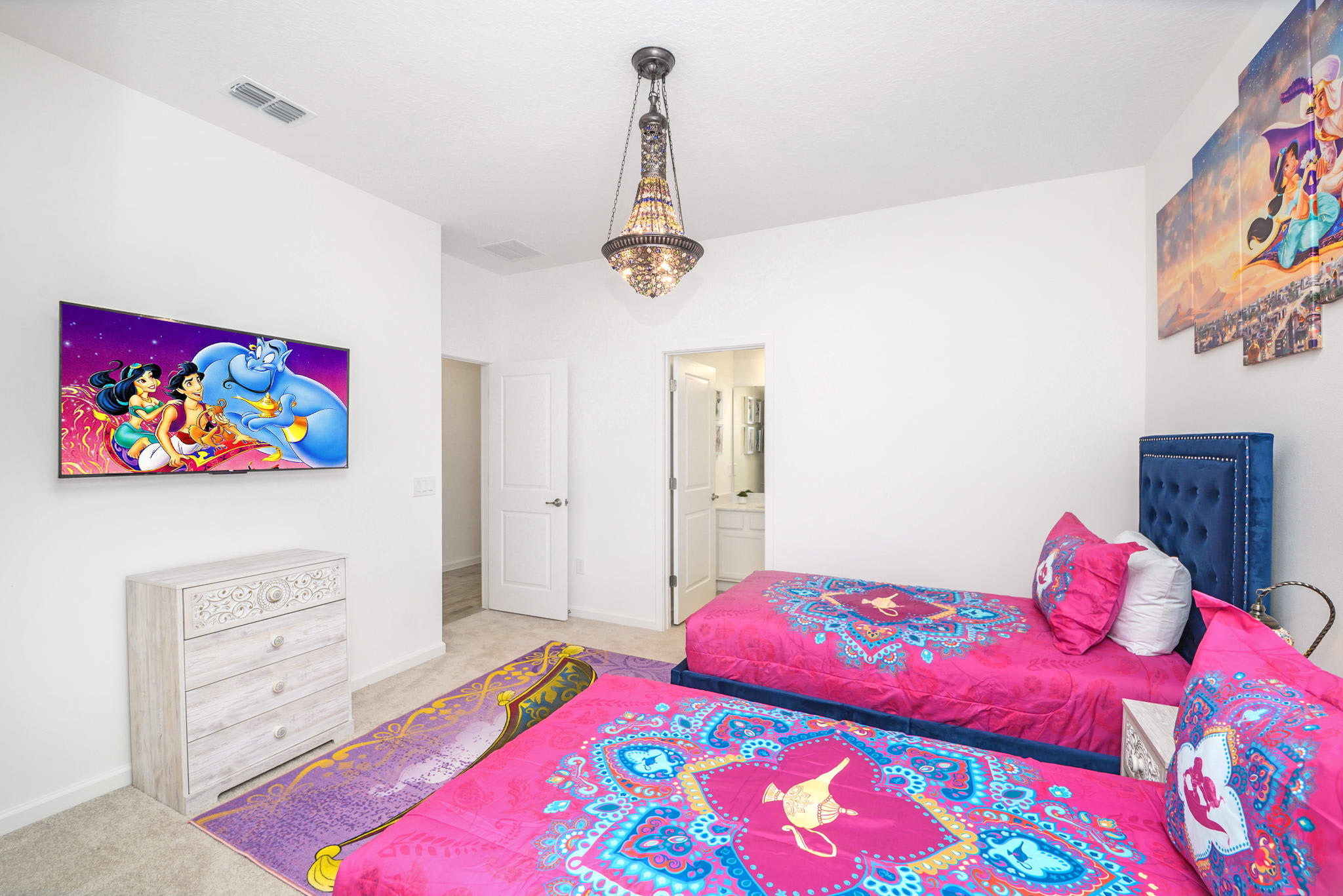 View of a themed bedroom with two twin beds and a white distressed dresser with a wall-mounted TV displaying an Aladdin scene. A large Aladdin-themed rug is on the carpeted floor, and an ornate chandelier hangs from the ceiling.