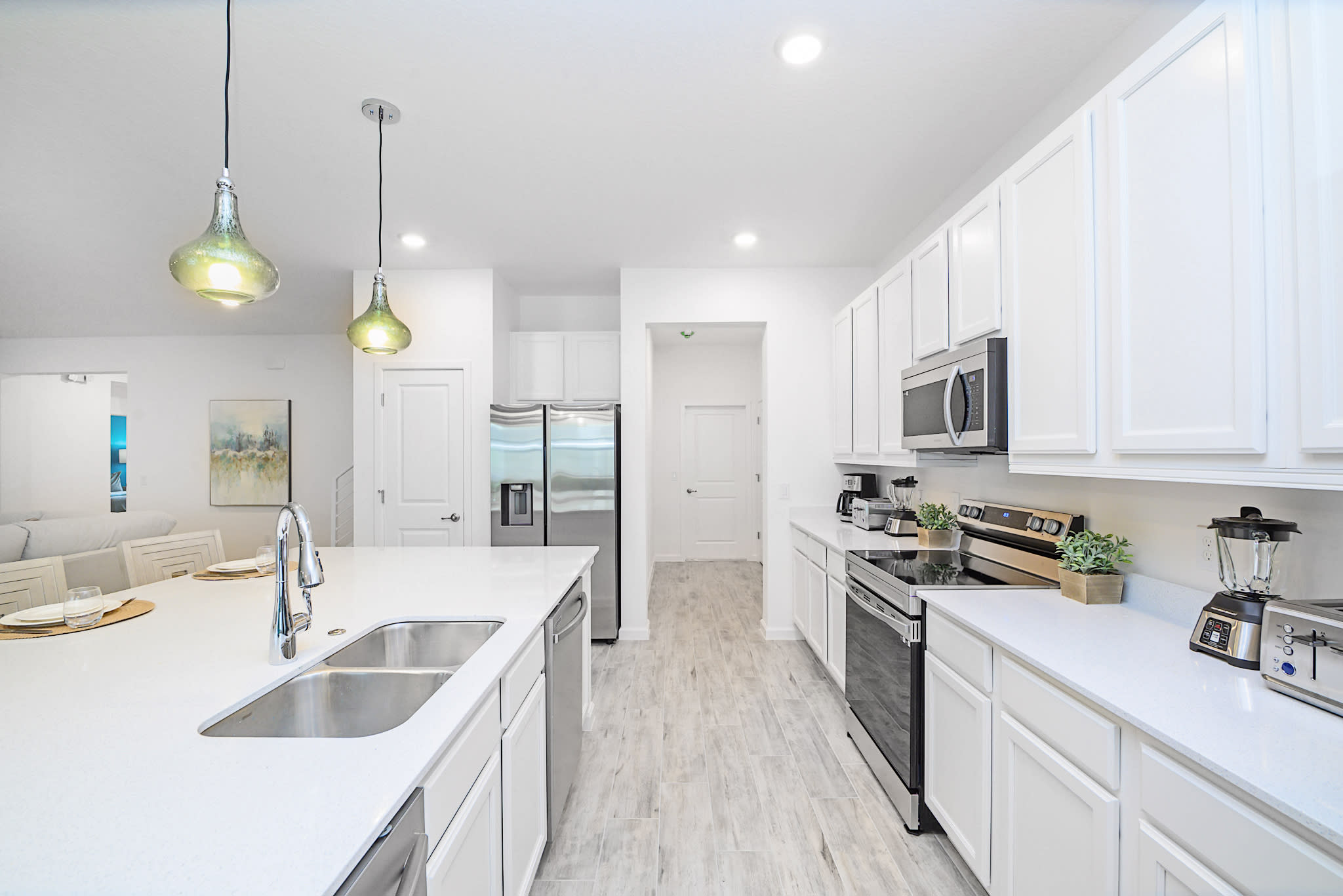 Bright and spacious galley kitchen featuring white shaker cabinets, stainless steel appliances, and light quartz countertops. Pendant lights hang above the island, and the open layout connects to the dining and living areas.