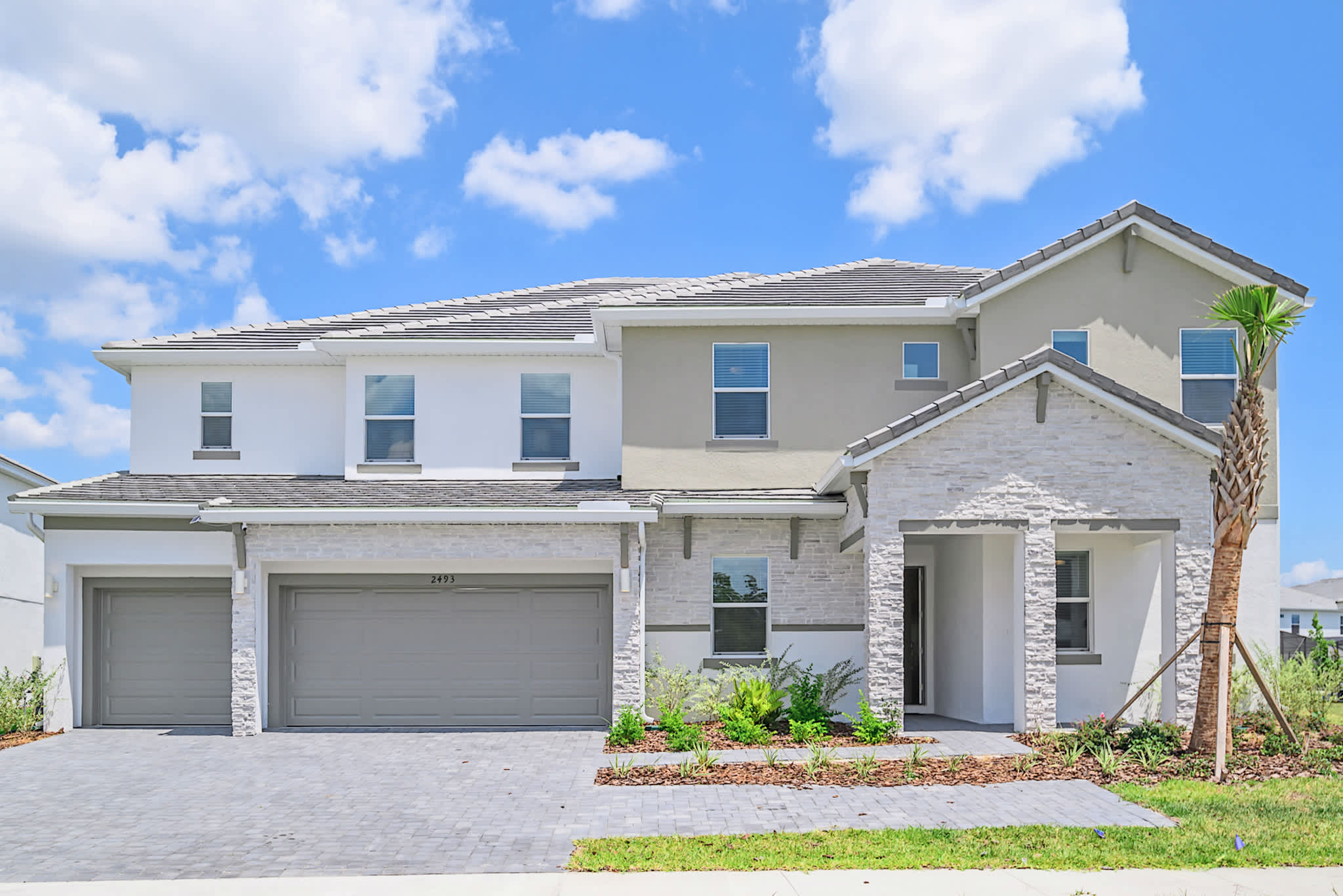 The modern two-story house exterior features a mix of white stucco and light gray stone cladding, with a gray tiled roof. A paved driveway leads to a two-car garage, and a small palm tree stands near the entrance.